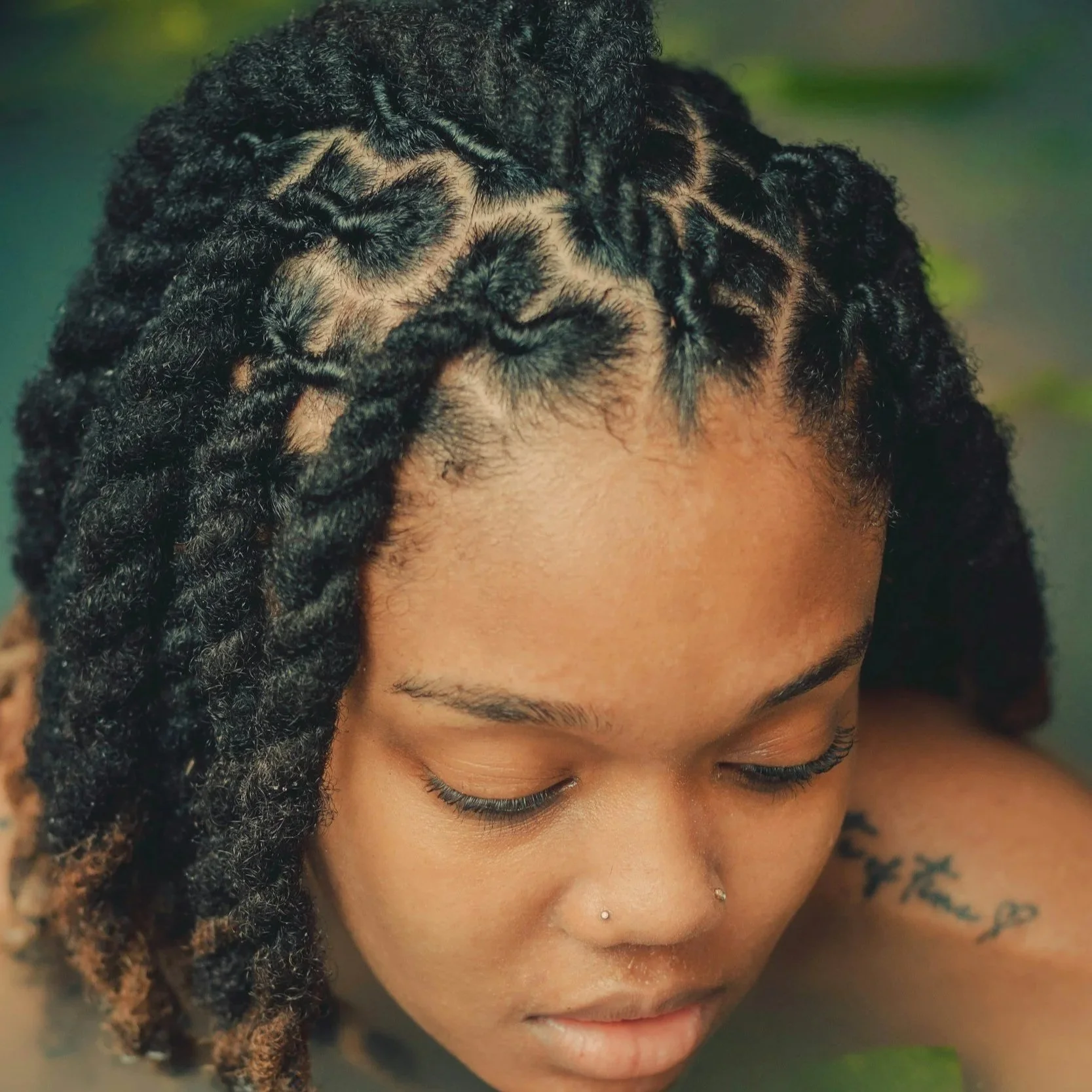 Close-up of a woman with dark, textured locs, her head tilted downward, showing her forehead, eyebrows, and closed eyes with long eyelashes.