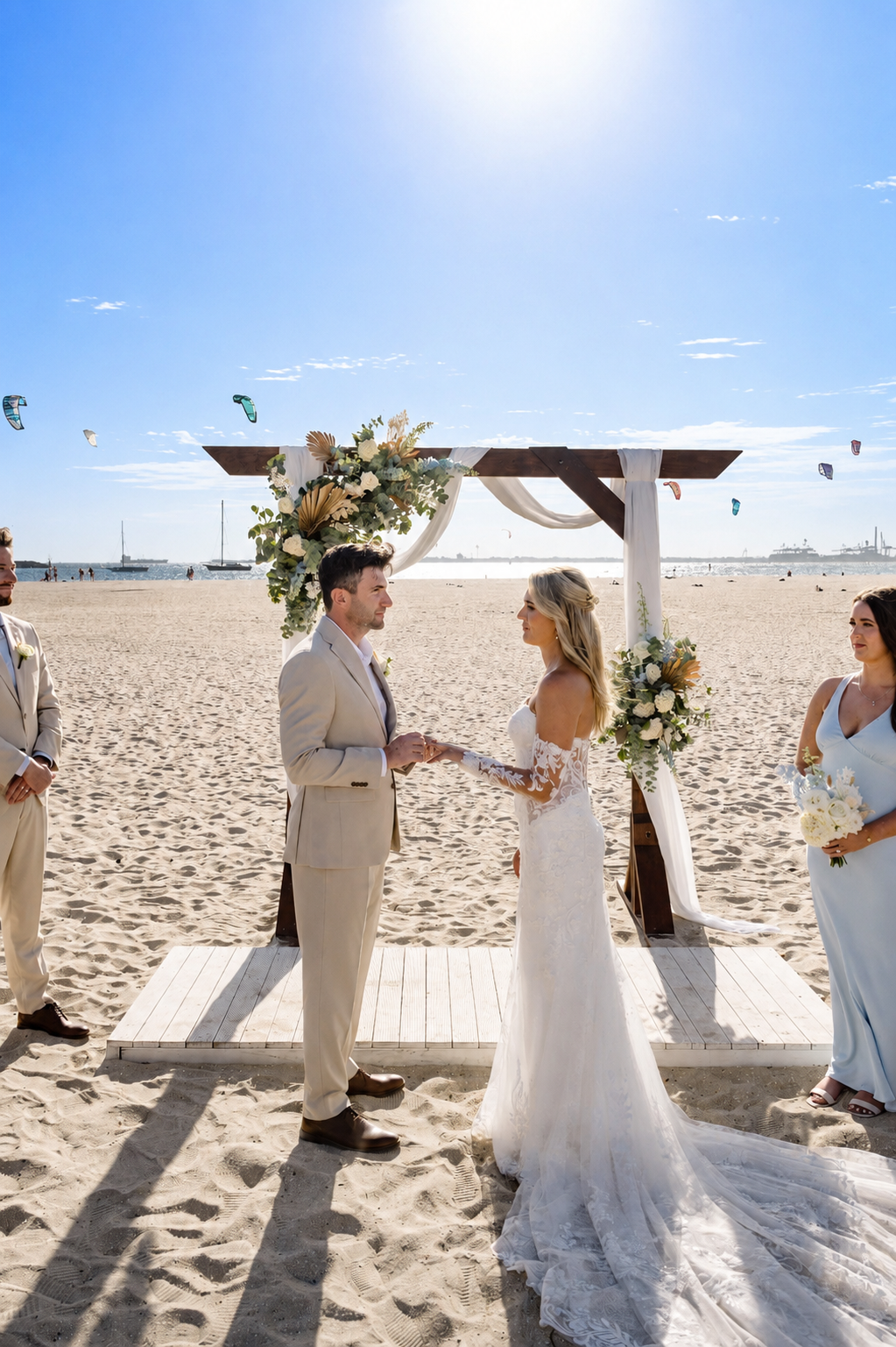 A beach wedding ceremony with a couple exchanging vows under a floral arch, surrounded by bridesmaids and groomsmen, with sailboats and kites in the background.