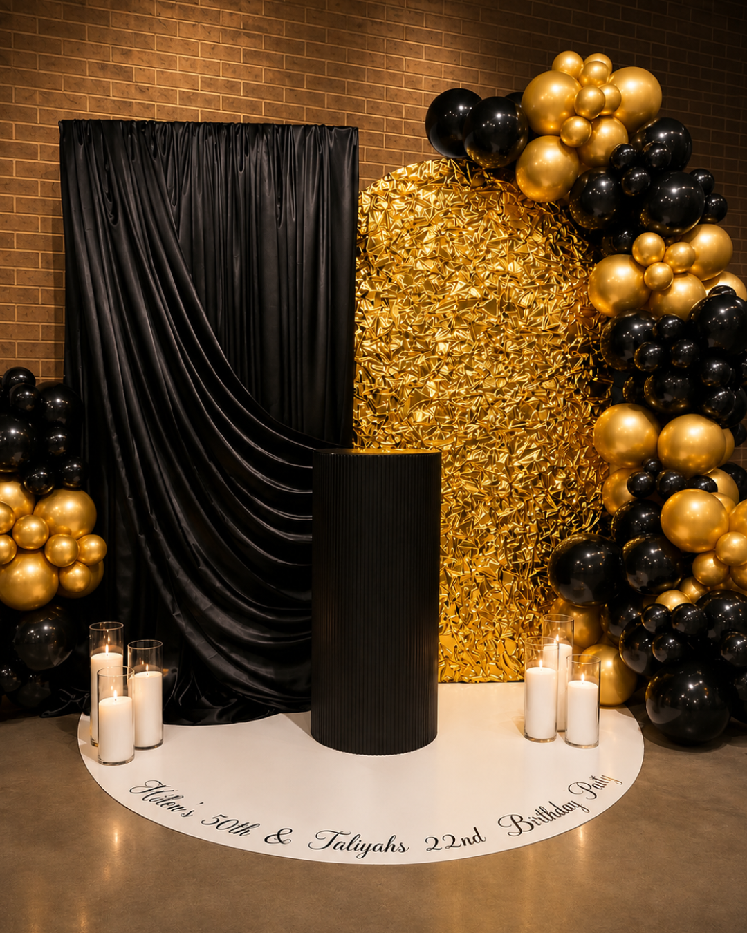 Elegant party backdrop with black and gold balloons, black draped curtain, black cylindrical column, white circular floor marker with personalized birthday message, and candles.