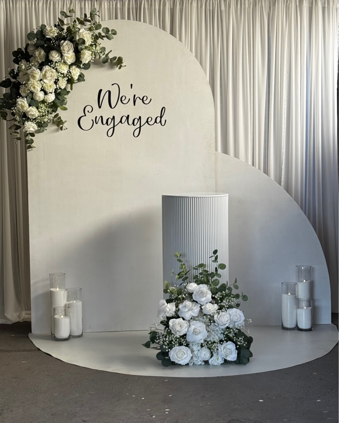 A wedding photo backdrop with white floral arrangements, candles in glass holders, and a sign that says 'We're Engaged' in black cursive.