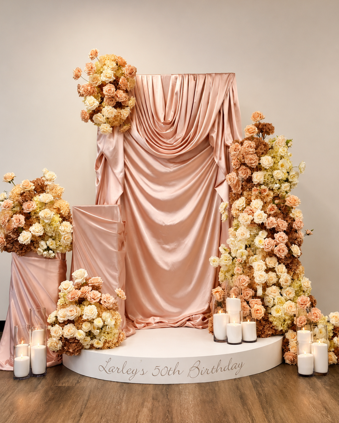 Elegant 50th birthday display with pink satin draping, peach and white roses, candles and a personalised “Larley’s 50th Birthday” sign.