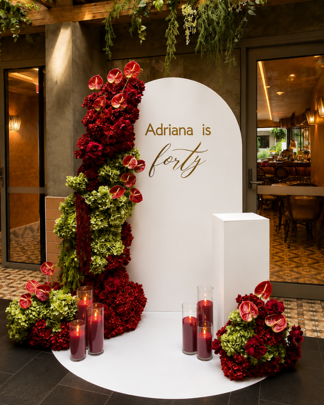 A decorative display with a white backdrop reading 'Adriana is forty,' adorned with red, green, and burgundy floral arrangements and candles in glass holders, at a restaurant or event venue entrance.
