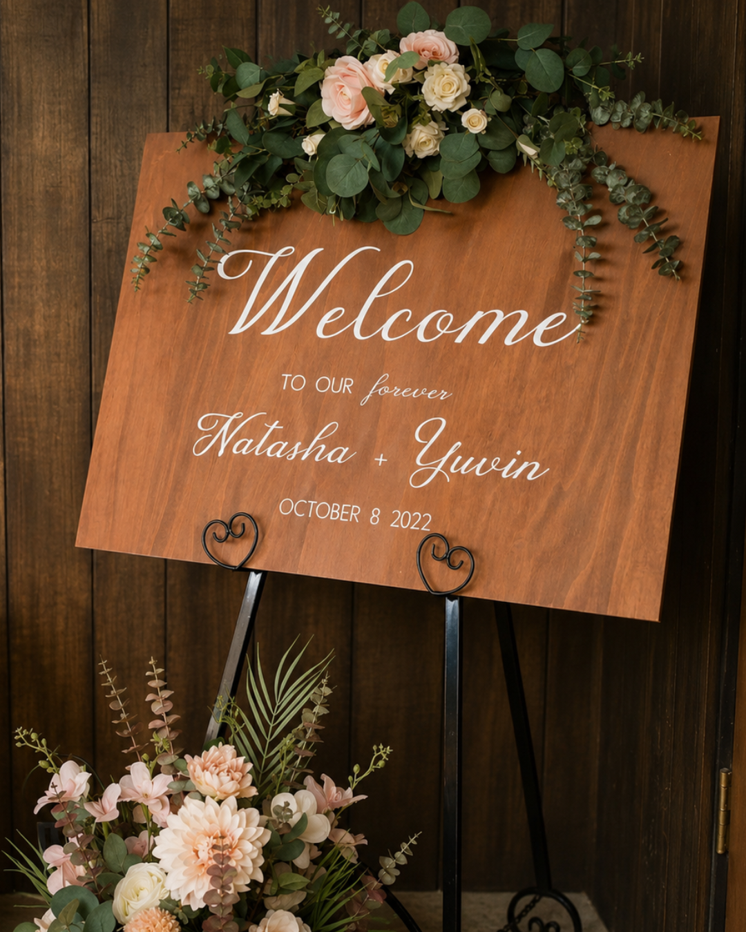 A wooden wedding welcome sign decorated with pink and white flowers and greenery, placed on a black metal stand.