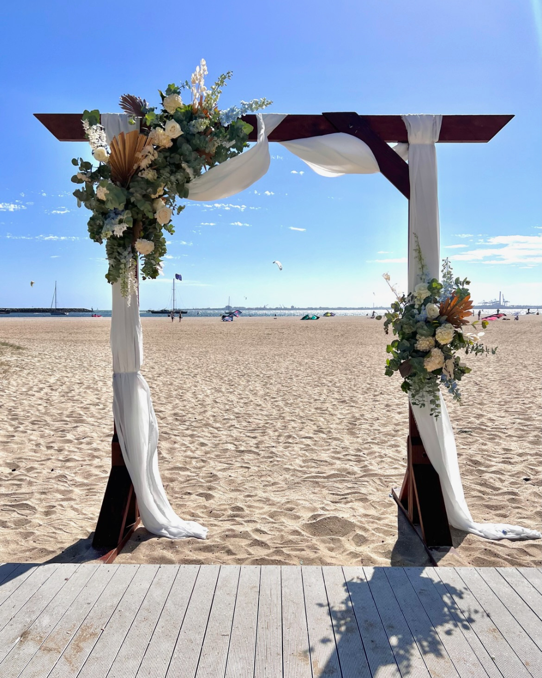 Beach wedding arch decorated with white and green flowers, fabric drapes, on a sandy beach with sailboats and kite surfers in the background under a blue sky.