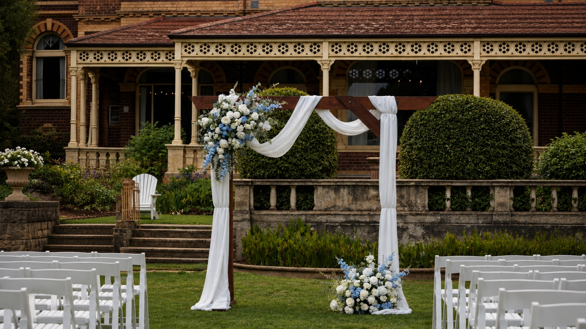 Wedding arch decorated with white and blue flowers and draped white fabric set in a lush garden with chairs for guests, in front of a historic brick house.