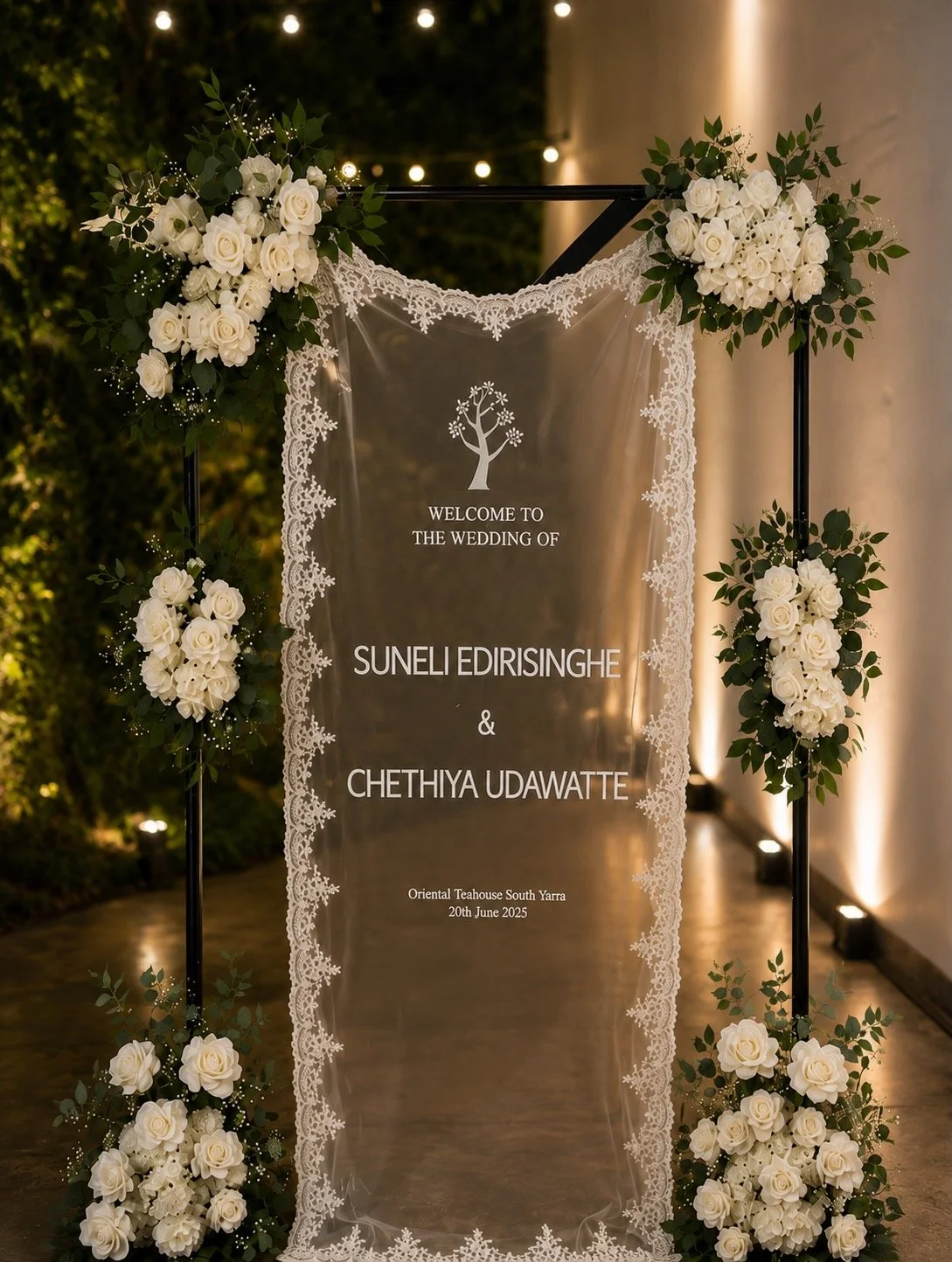 Wedding welcome sign on sheer fabric, decorated with white roses and greenery in an elegant outdoor setting.