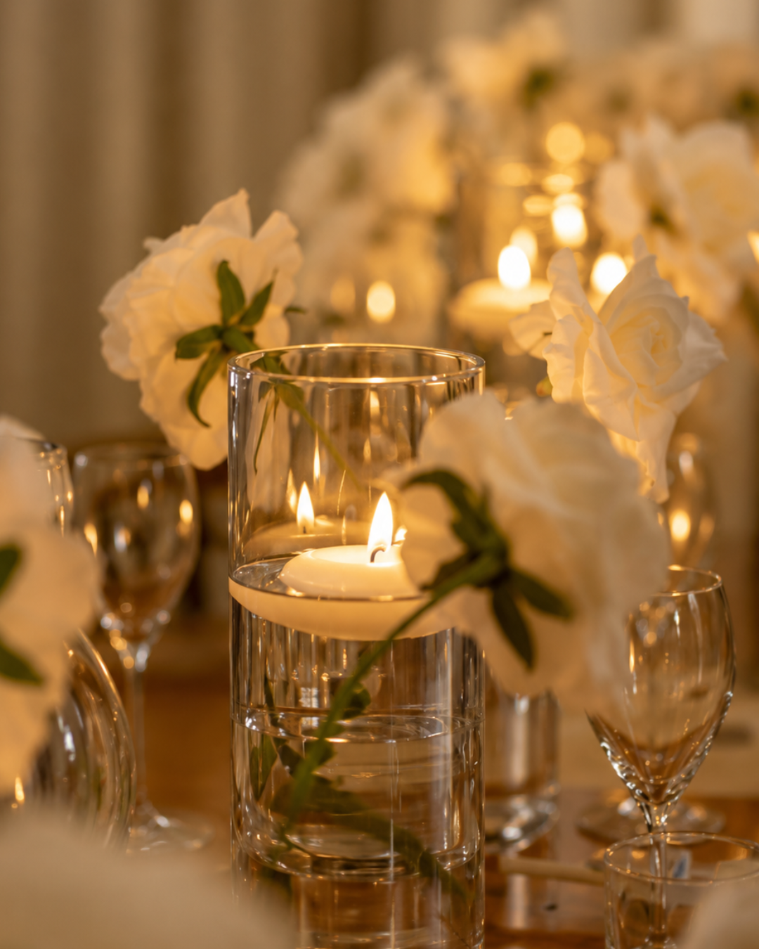 A close-up of a table setting with a glass candle holder containing a lit candle, surrounded by white flowers and wine glasses.