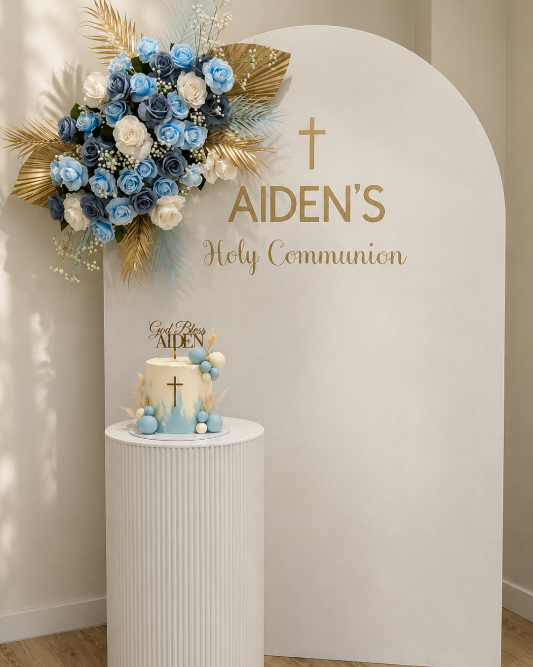 A white sign with gold lettering that reads "Aiden's Holy Communion" and a gold cross above the name. Decorated with a large floral arrangement of blue, white, and gold leaves. A small table with a white and blue cake featuring a gold cross and a cake topper that says "God Bless Aiden," surrounded by blue and white spherical decorations.