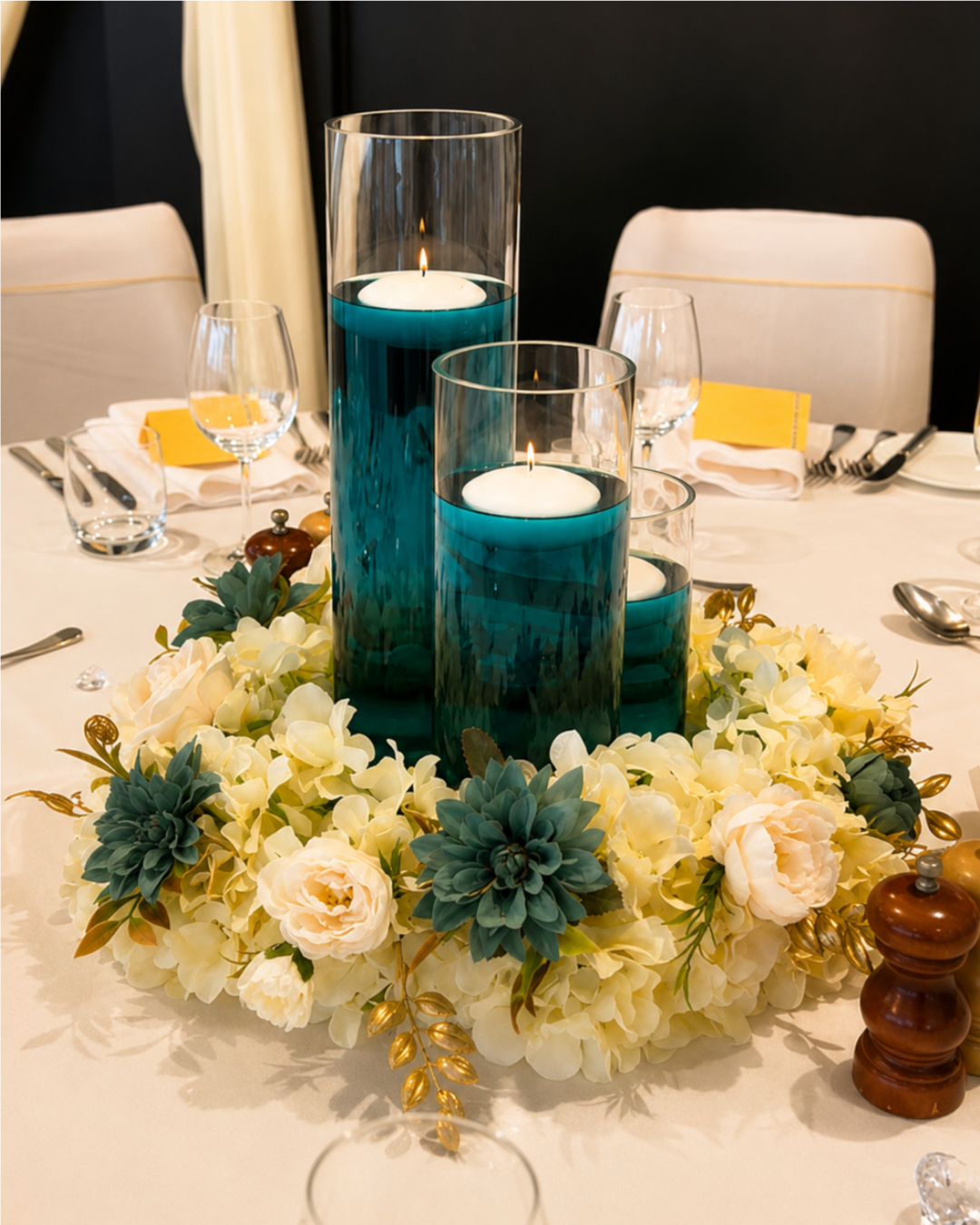 Elegant dining table centerpiece with three tall blue glass candle holders, white floating candles, surrounded by white and green floral arrangements, set for a formal dinner.