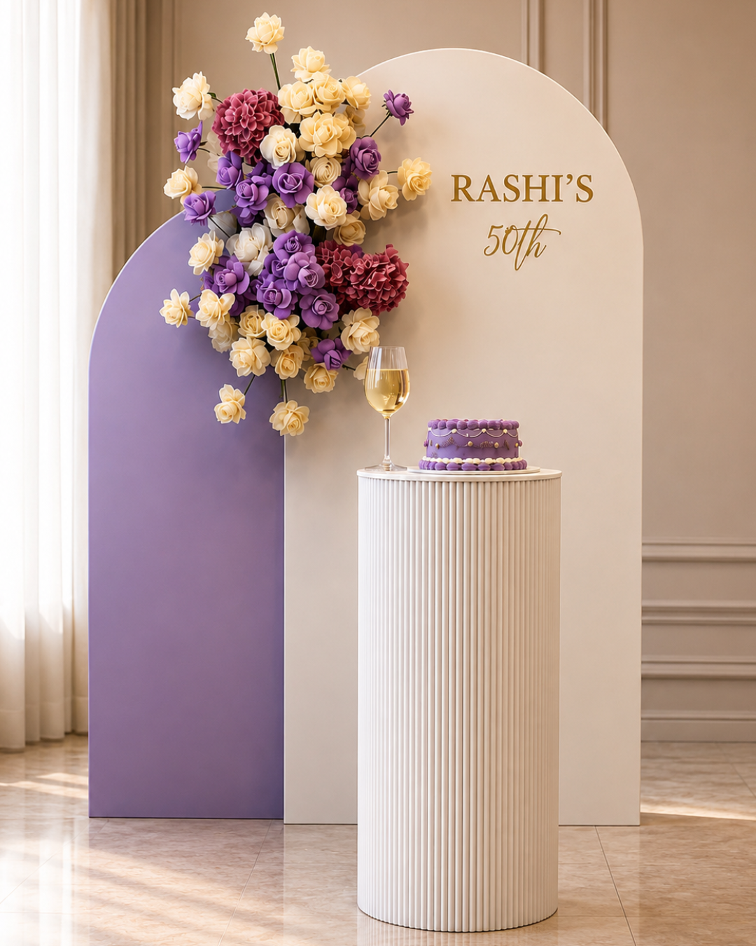 Elegant birthday celebration setup with a purple and white floral backdrop, a cake on a ribbed column, a glass of champagne, and a sign reading 'RASHI'S 50th'.