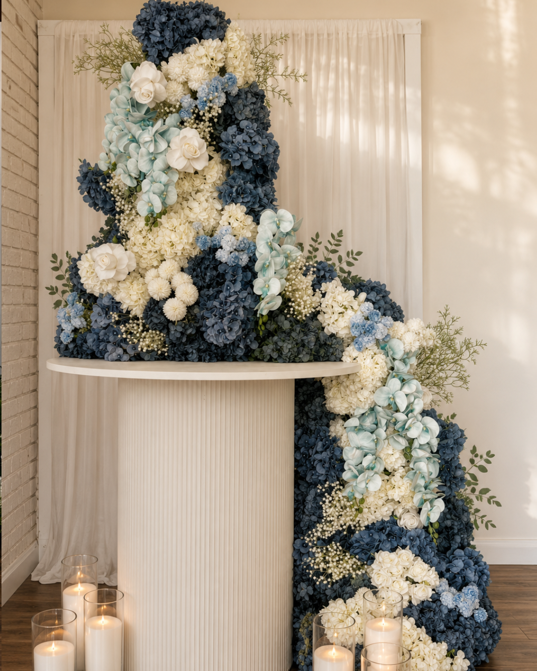 Elegant floral arrangement with white, blue, and cream flowers cascading from a white semi-circular table, with lit candles at the base, set against a backdrop of white curtains and a brick wall.