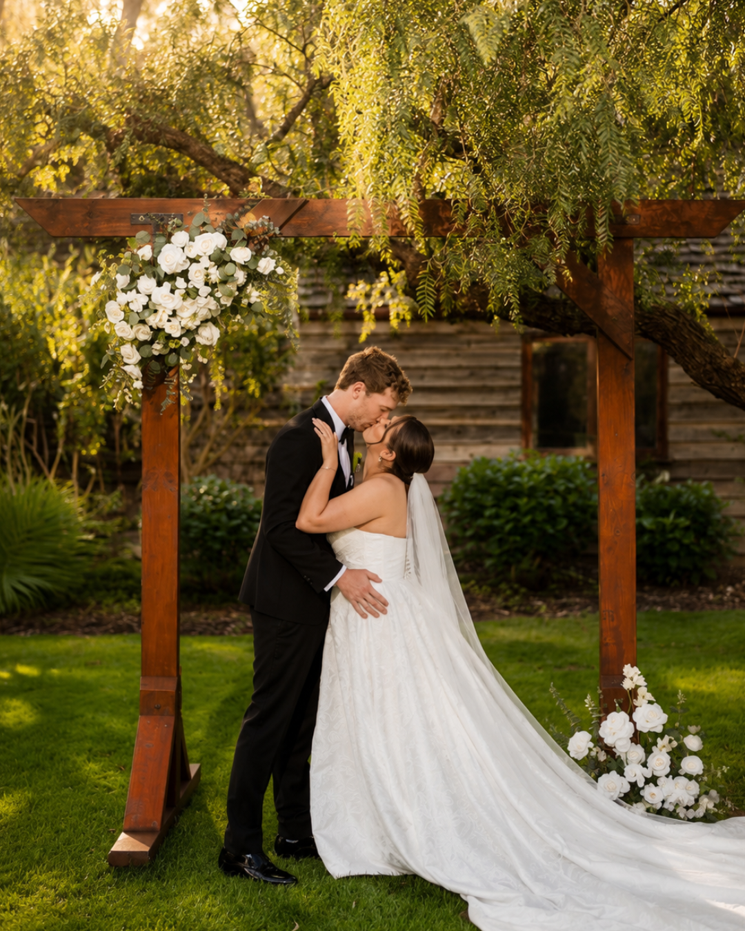 Couple kissing at their outdoor wedding ceremony with a wooden arch decorated with white flowers.