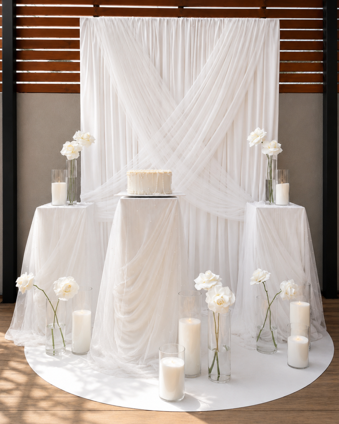 Elegant wedding or event setup with a white table and fabric backdrop, decorated with white roses in glass vases and candles in tall glass holders.