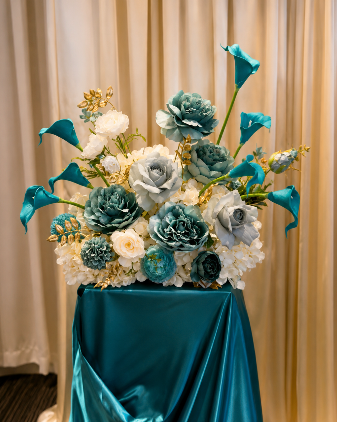 Blue and white floral arrangement with roses, calla lilies, and hydrangeas on a satin-covered table.