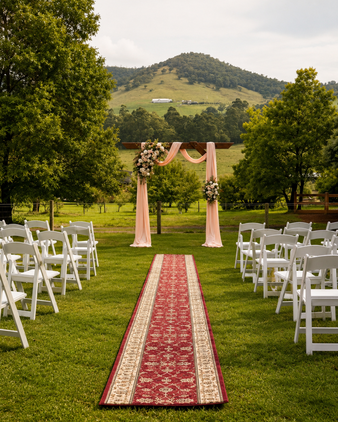 Outdoor wedding ceremony setup with white chairs arranged on sides of a red and beige aisle rug leading to a pink-draped wooden arch decorated with floral arrangements, set on a grassy field with trees and a mountain in the background.