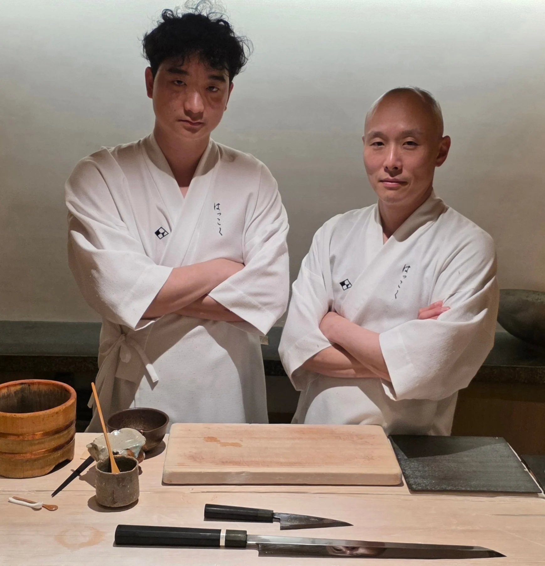 Two men dressed in traditional Japanese chef uniforms standing behind a wooden counter in a kitchen, with utensils, bowls, and a cutting board in front of them.