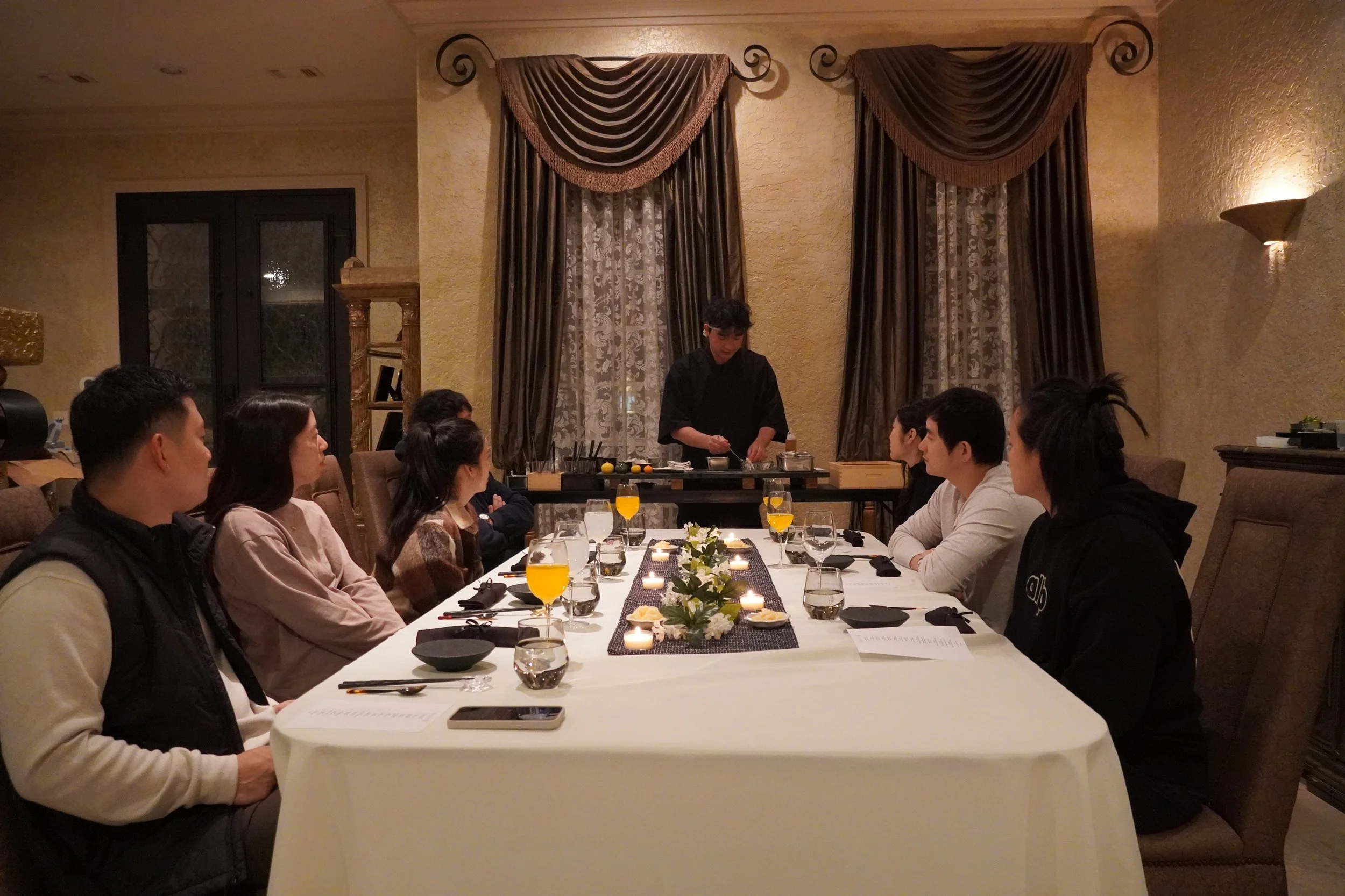 A group of people seated around a long, elegant dining table in a dimly lit room with dark curtains, raising glasses in a toast, while a chef prepares food at the table's end.