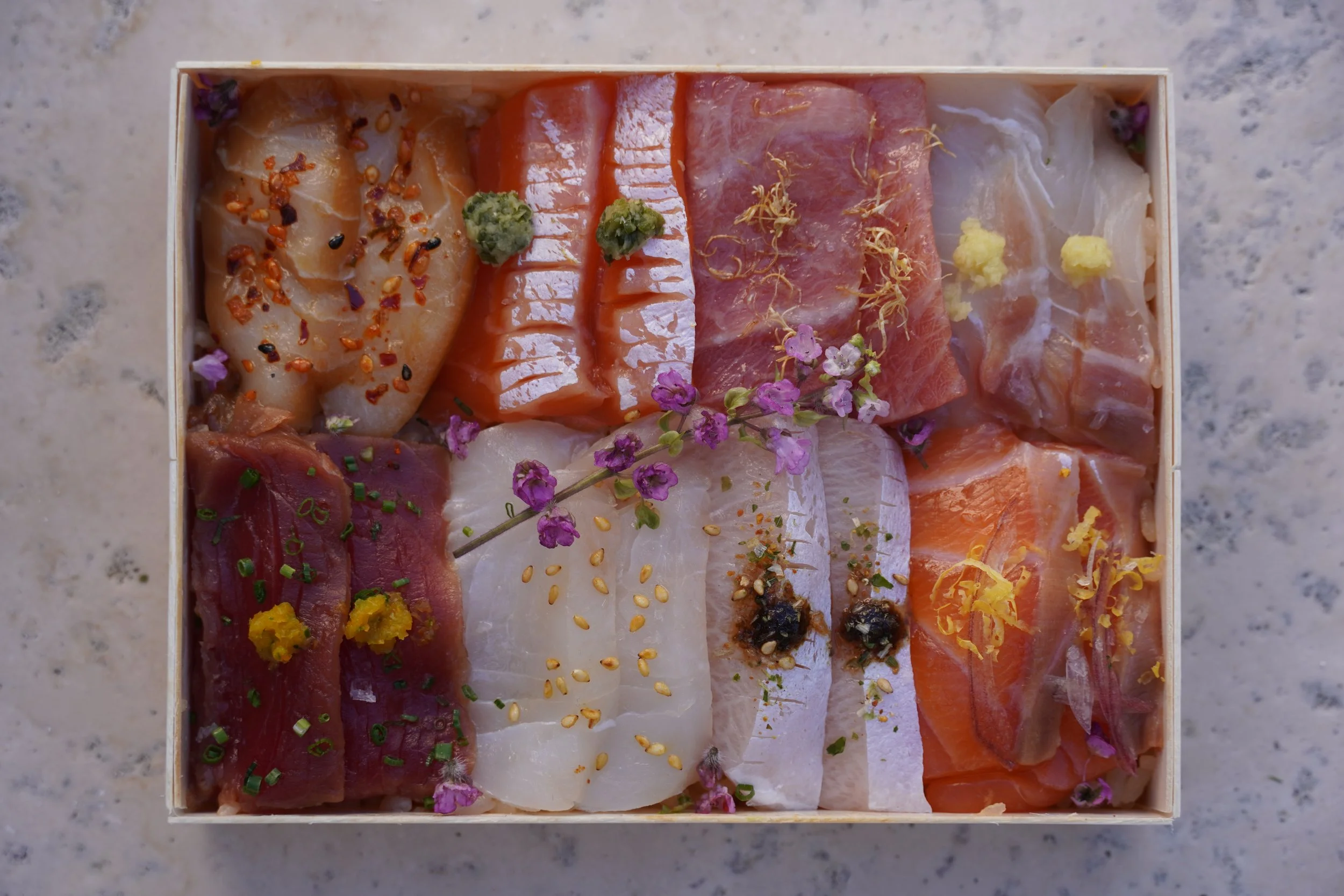 Assorted slices of raw fish and seafood arranged in a wooden box, garnished with herbs, spices, and edible flowers.