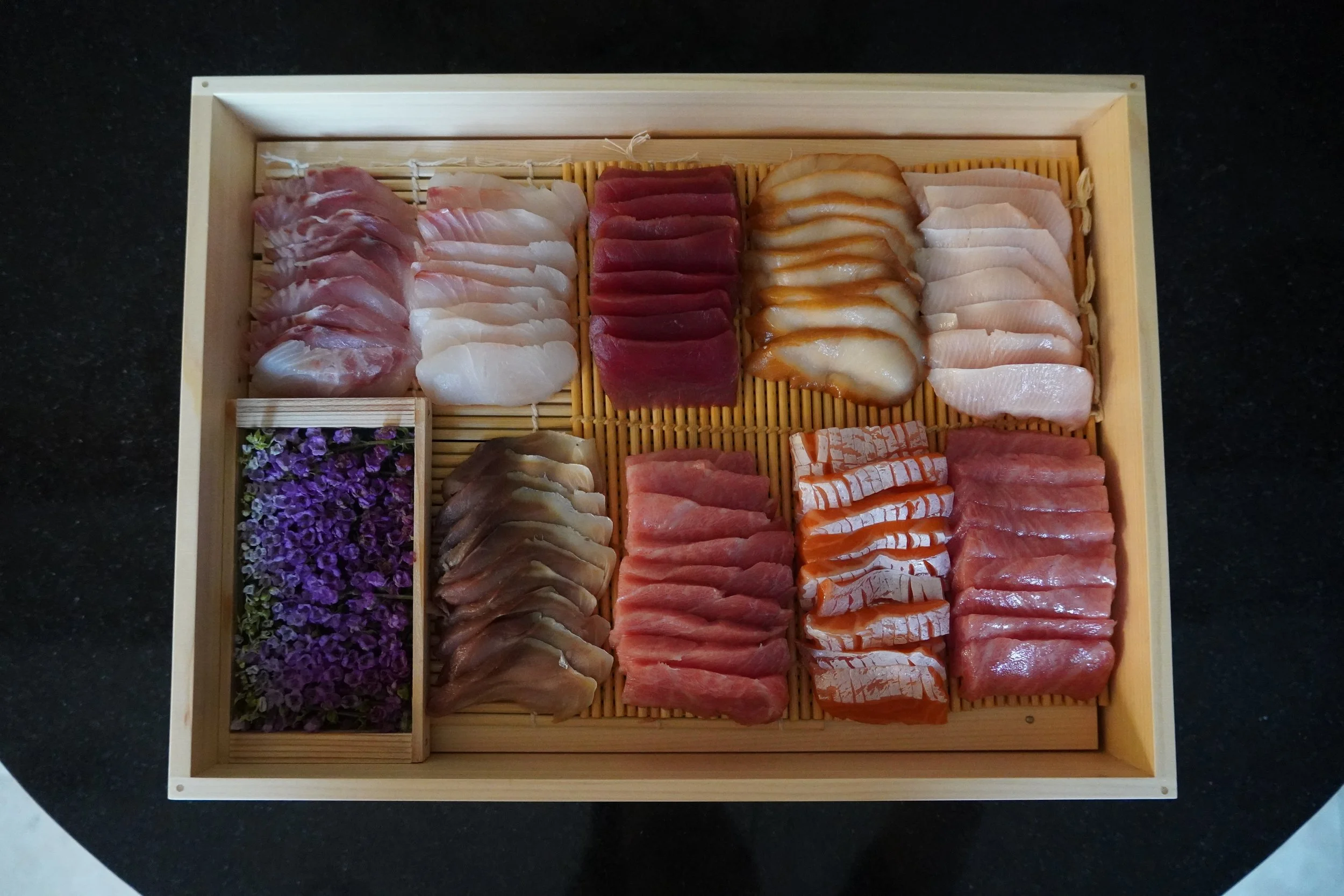 Assorted slices of sashimi arranged on a wooden tray with decorative flowers in a small section.