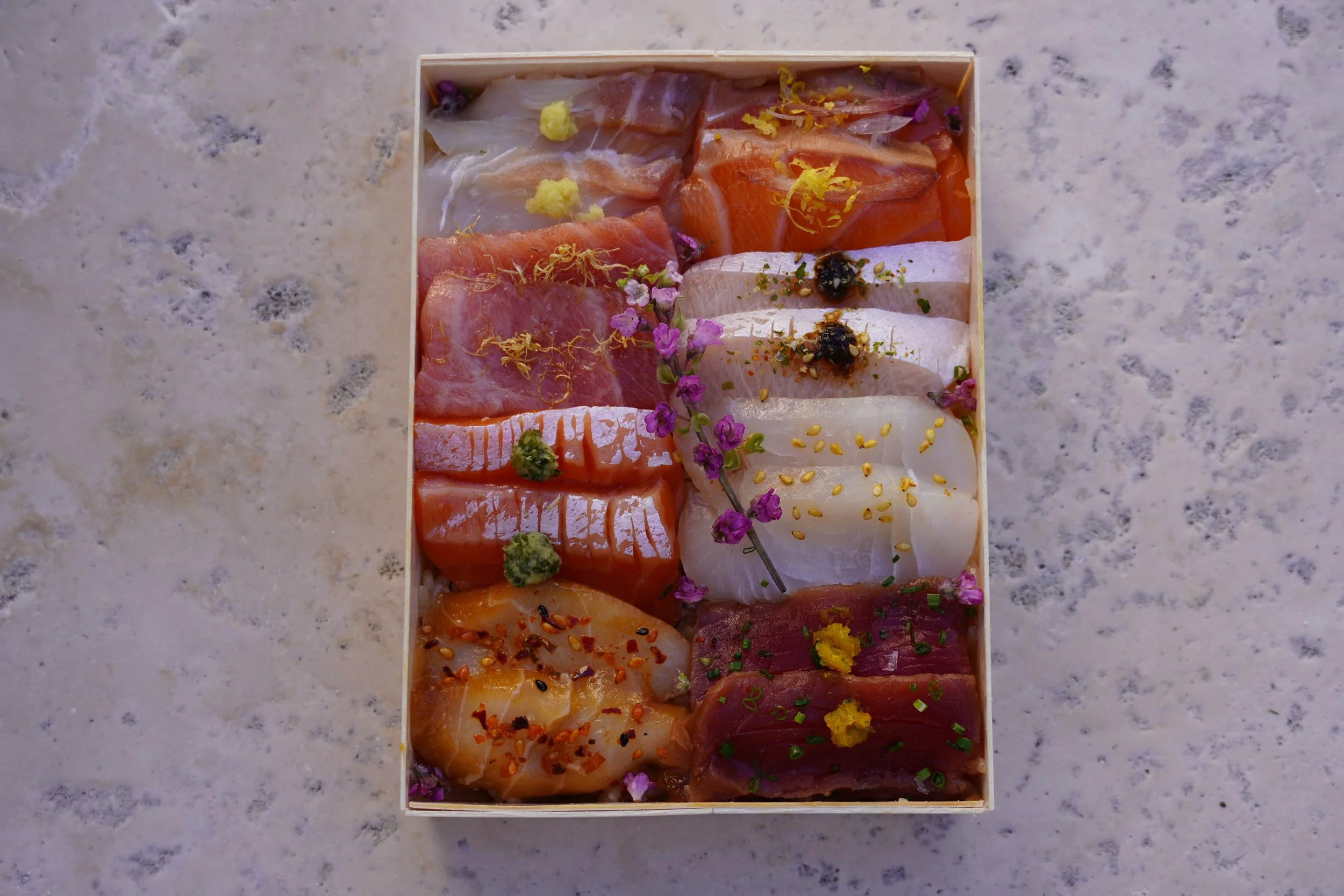 Assorted slices of sashimi arranged in a box, garnished with flowers, wasabi, and various seasonings on a marble surface.