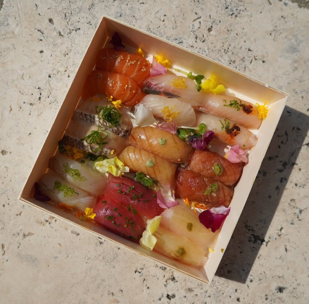 Assorted slices of sashimi garnished with edible flowers and green onions in a white box.
