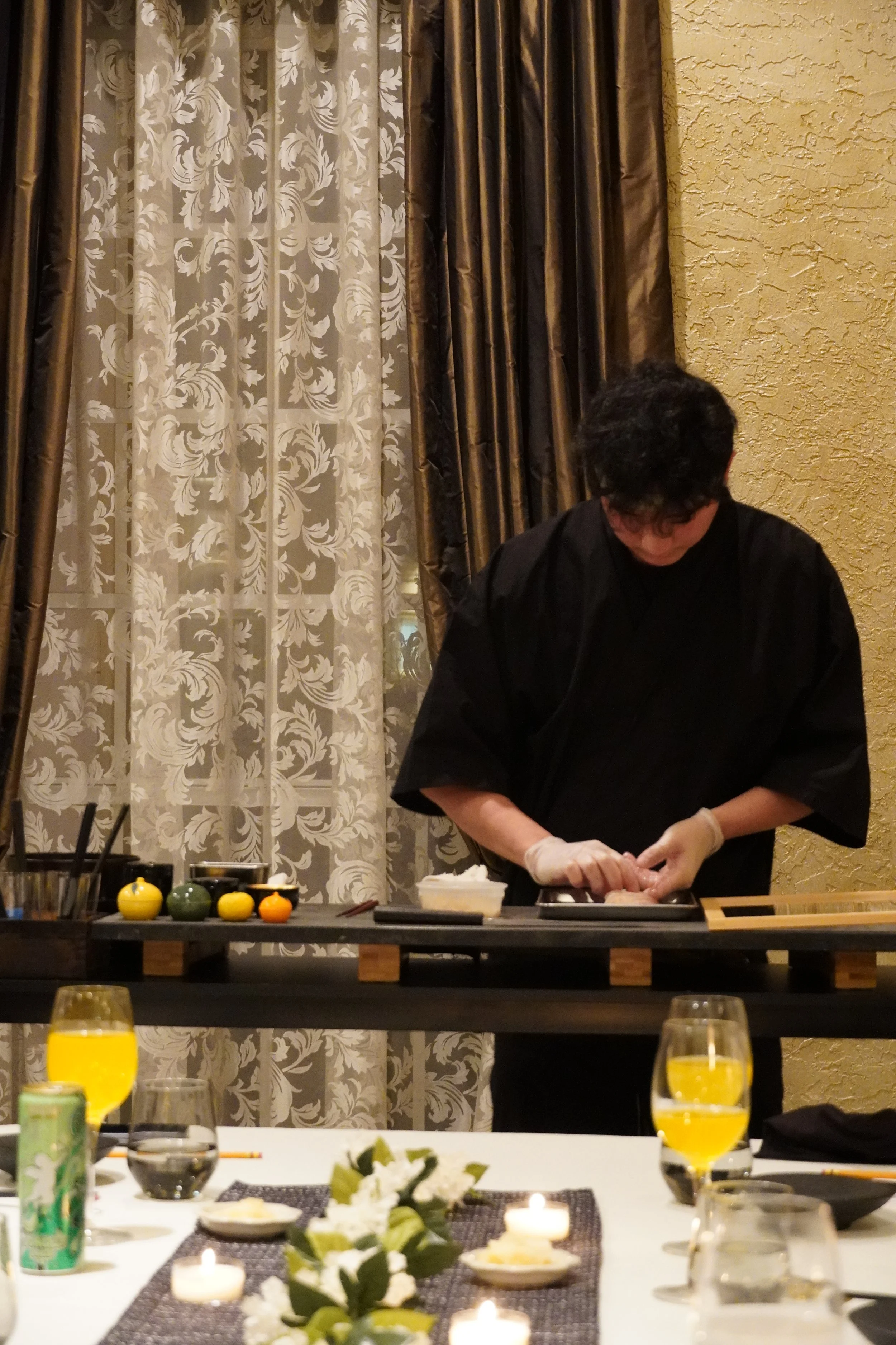A person wearing a black shirt and gloves prepares sushi at a table inside a restaurant, with decorative curtains and textured yellow wall in the background.
