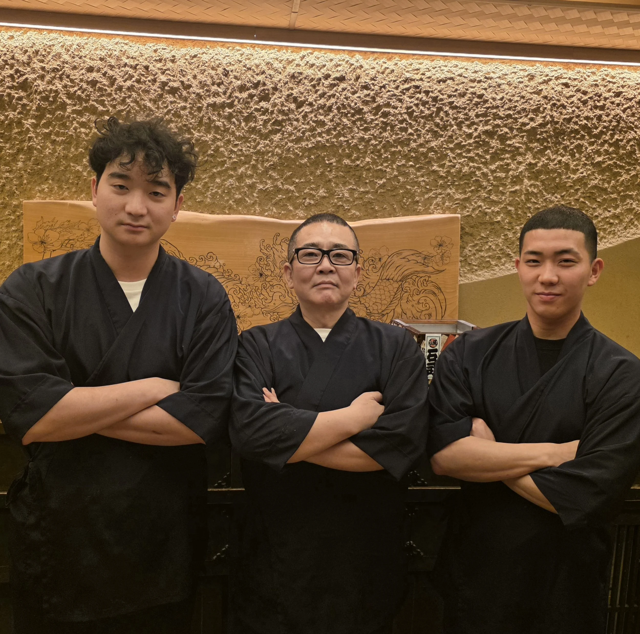 Three men wearing black traditional martial arts uniforms standing in front of a textured beige wall with a piece of wooden art featuring a dragon design.