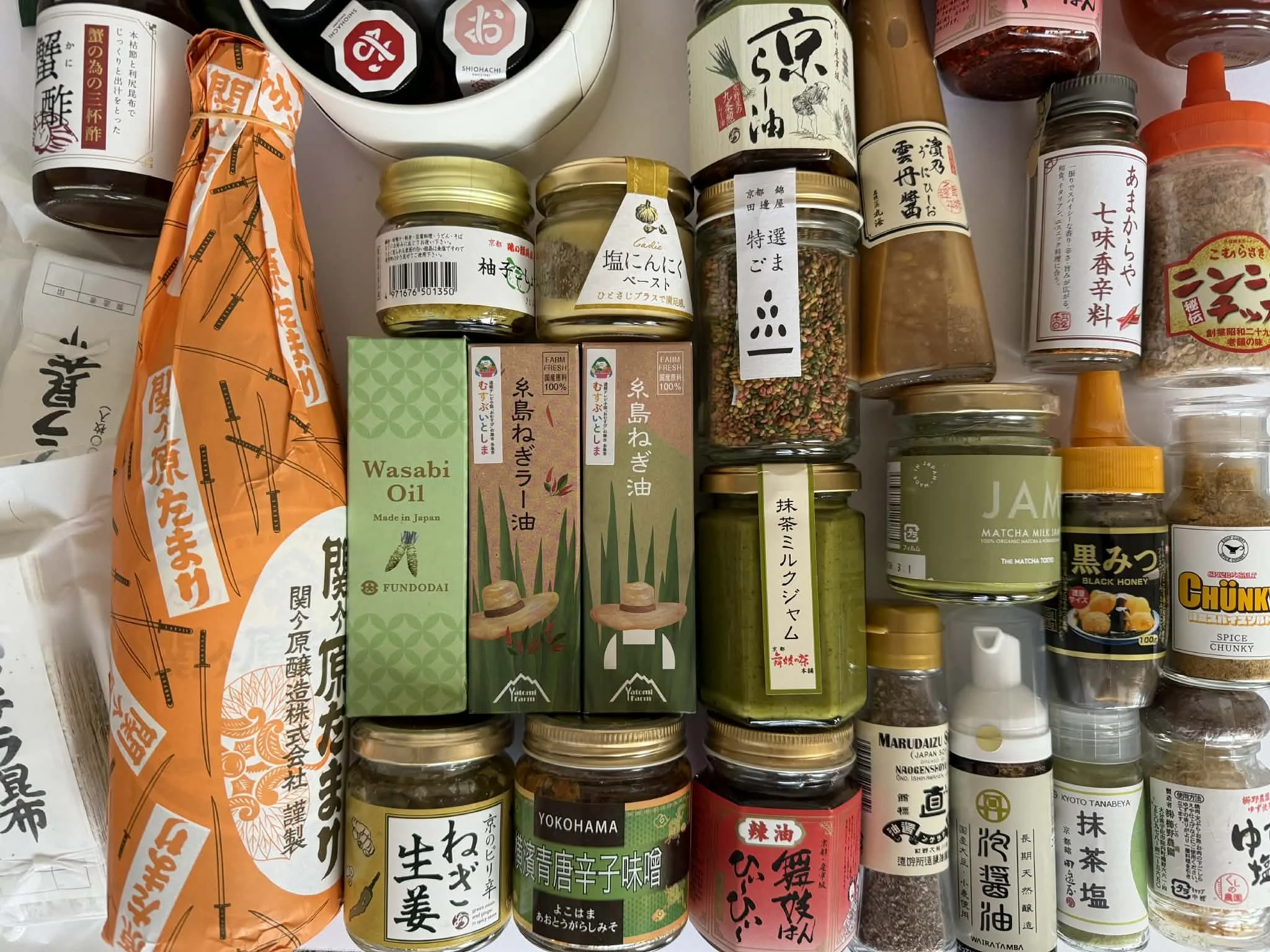 A collection of various Japanese food condiments and sauces, including soy sauce, wasabi oil, sesame seeds, and seasoned spices, in jars and bottles on a shelf.