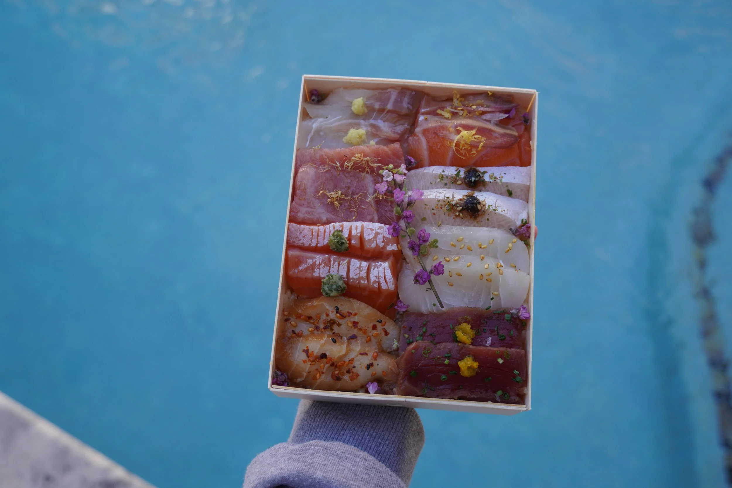 Hand holding a wooden box of assorted sashimi slices decorated with herbs and edible flowers, over a blue surface.