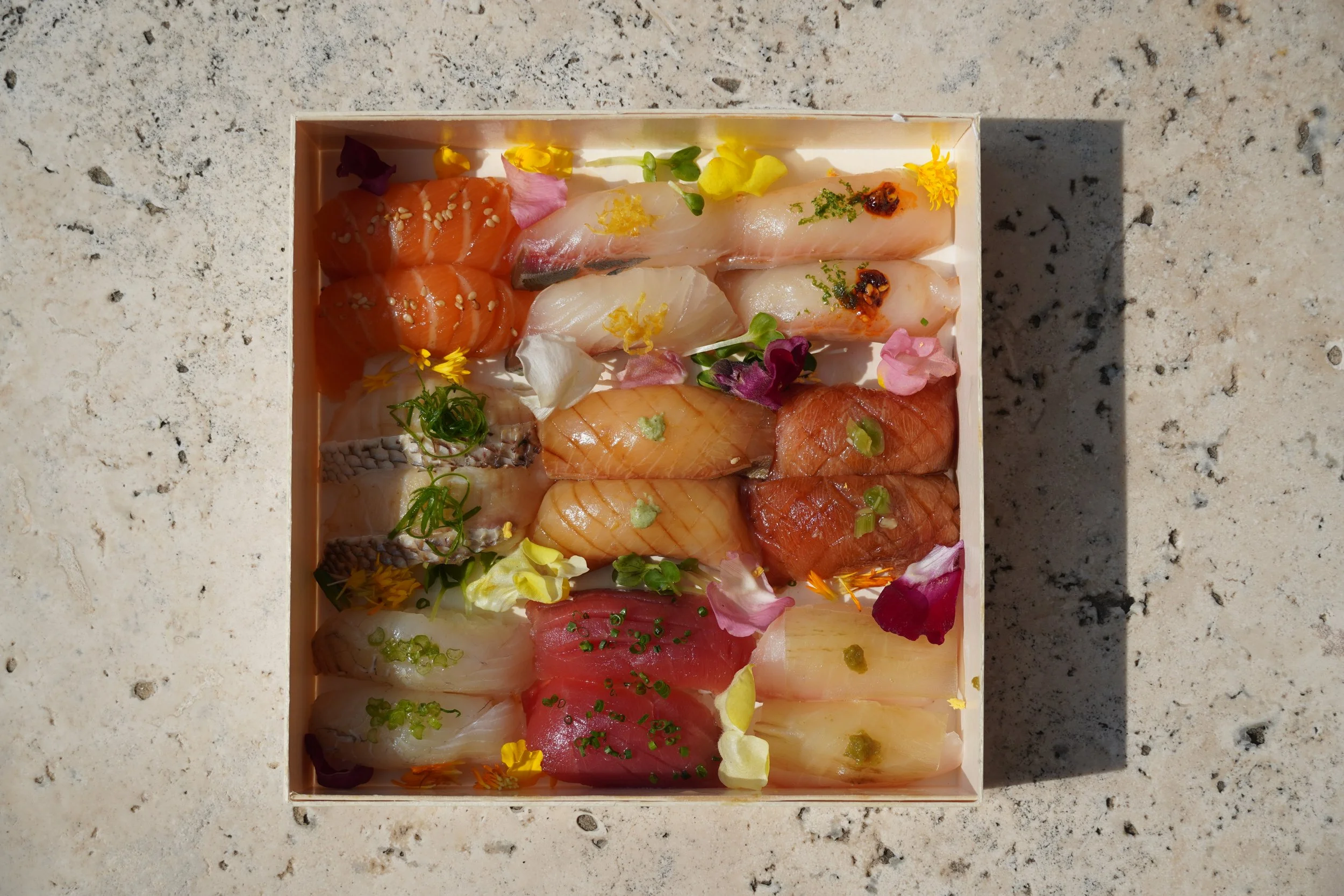 Assorted slices of sashimi arranged in a box, garnished with edible flowers and herbs, on a light-colored stone surface.