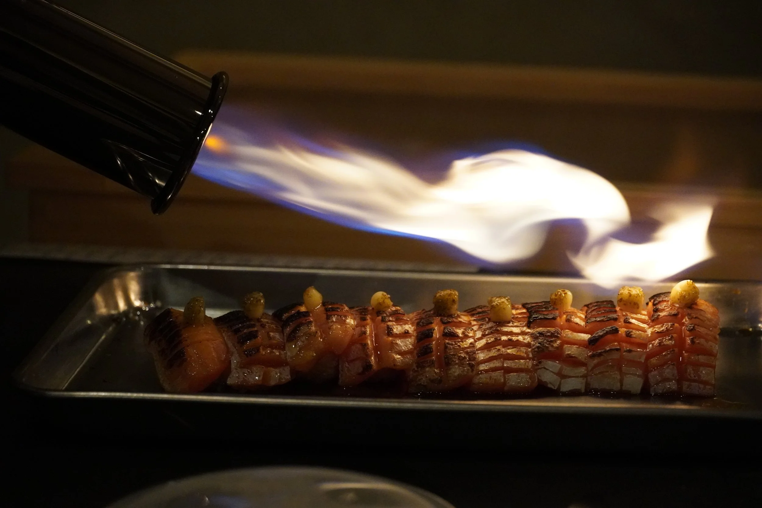 A close-up view of a commercial kitchen oven with a flame coming out of a gas burner. The oven contains a row of skewered food items, possibly meat and vegetables, being cooked over the flame.