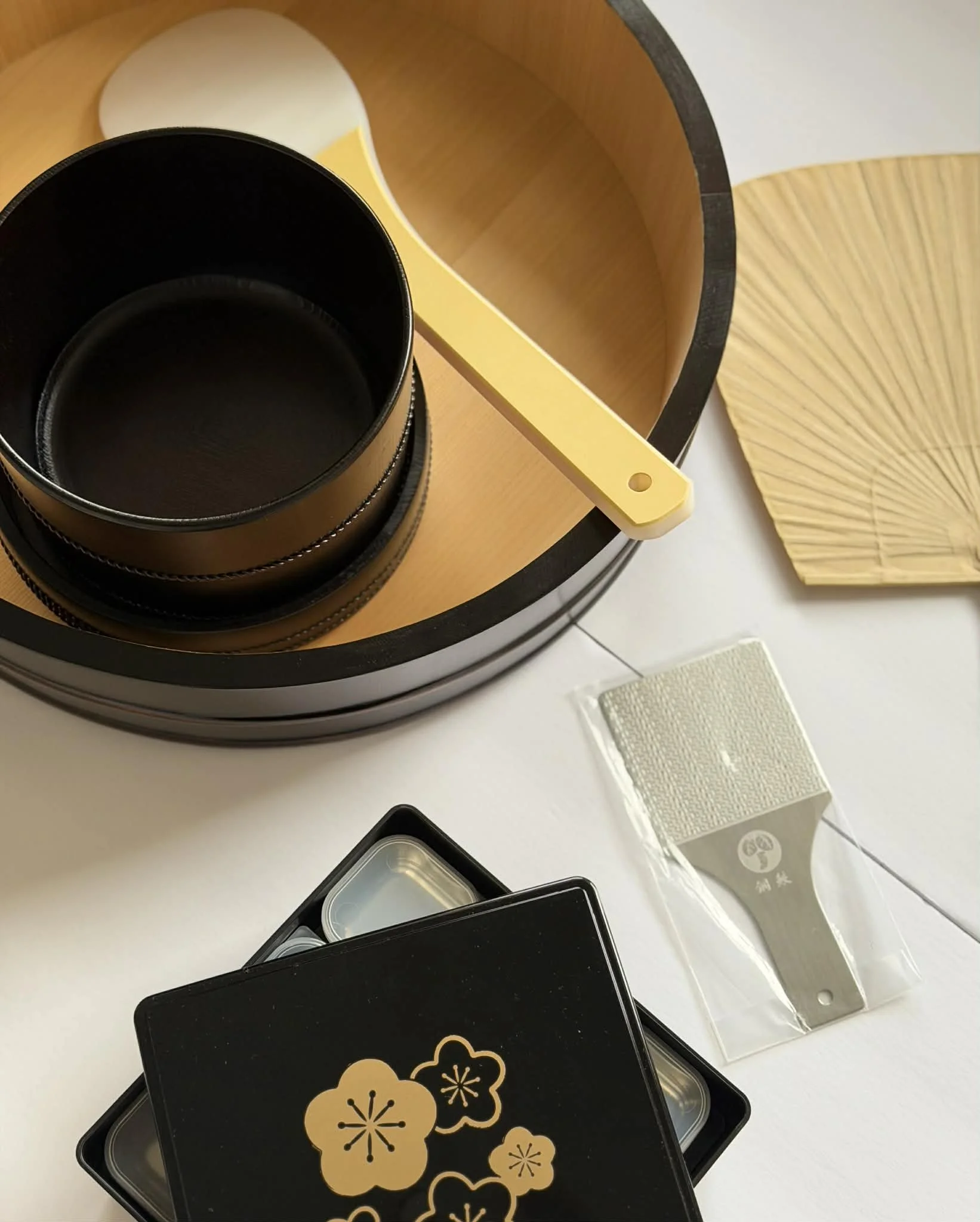 A black box with spring or cherry blossom motifs on the lid, open to reveal empty compartments, placed on a white table alongside a small plastic container and a beige rectangular box with Japanese characters. In the background, there's a decorative 