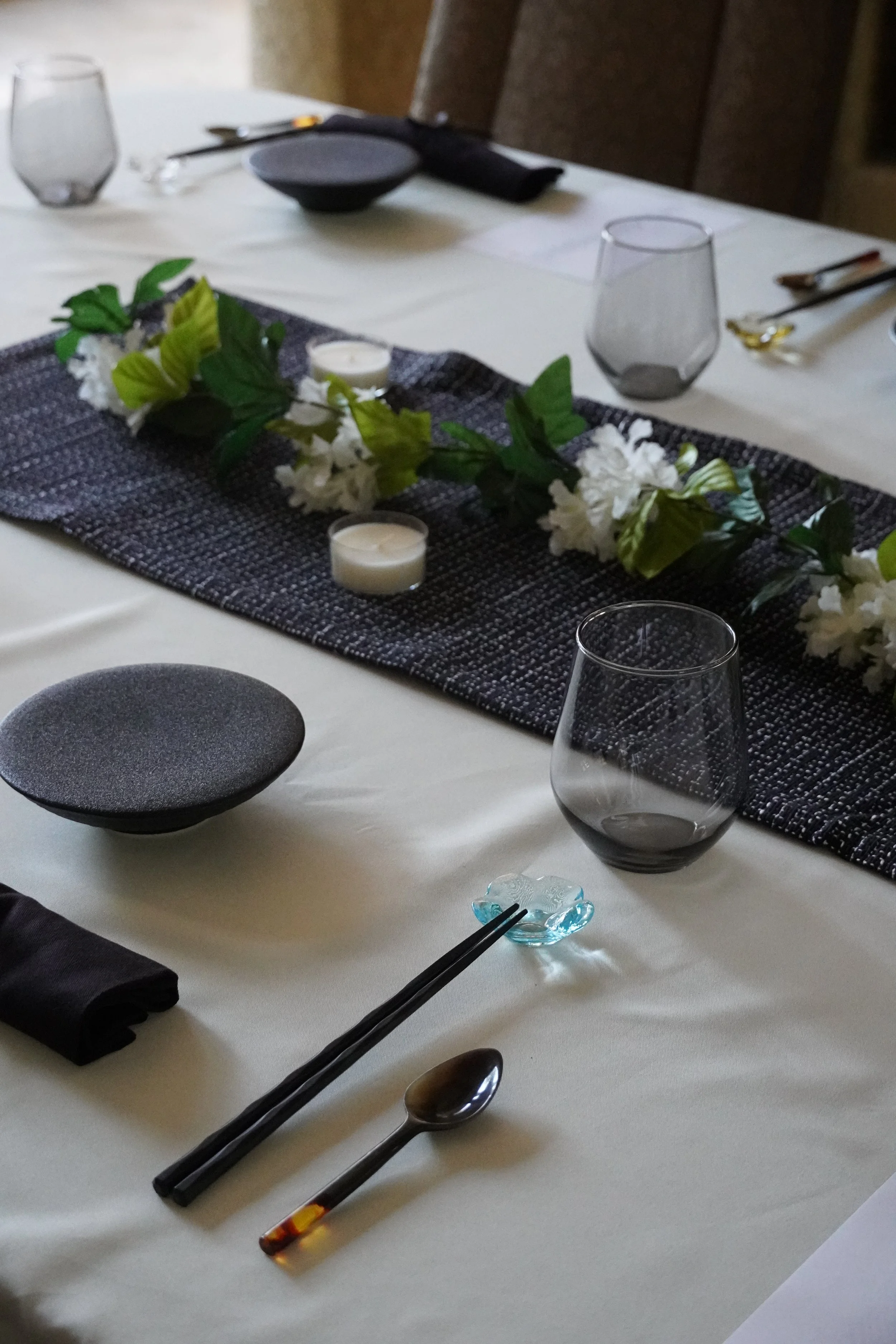 A dining table set for a meal with black chopsticks, a spoon, and a small blue decorative dish in the foreground. There is a black textured plate runner with white flowers and green leaves, candles, and empty glasses on the table.