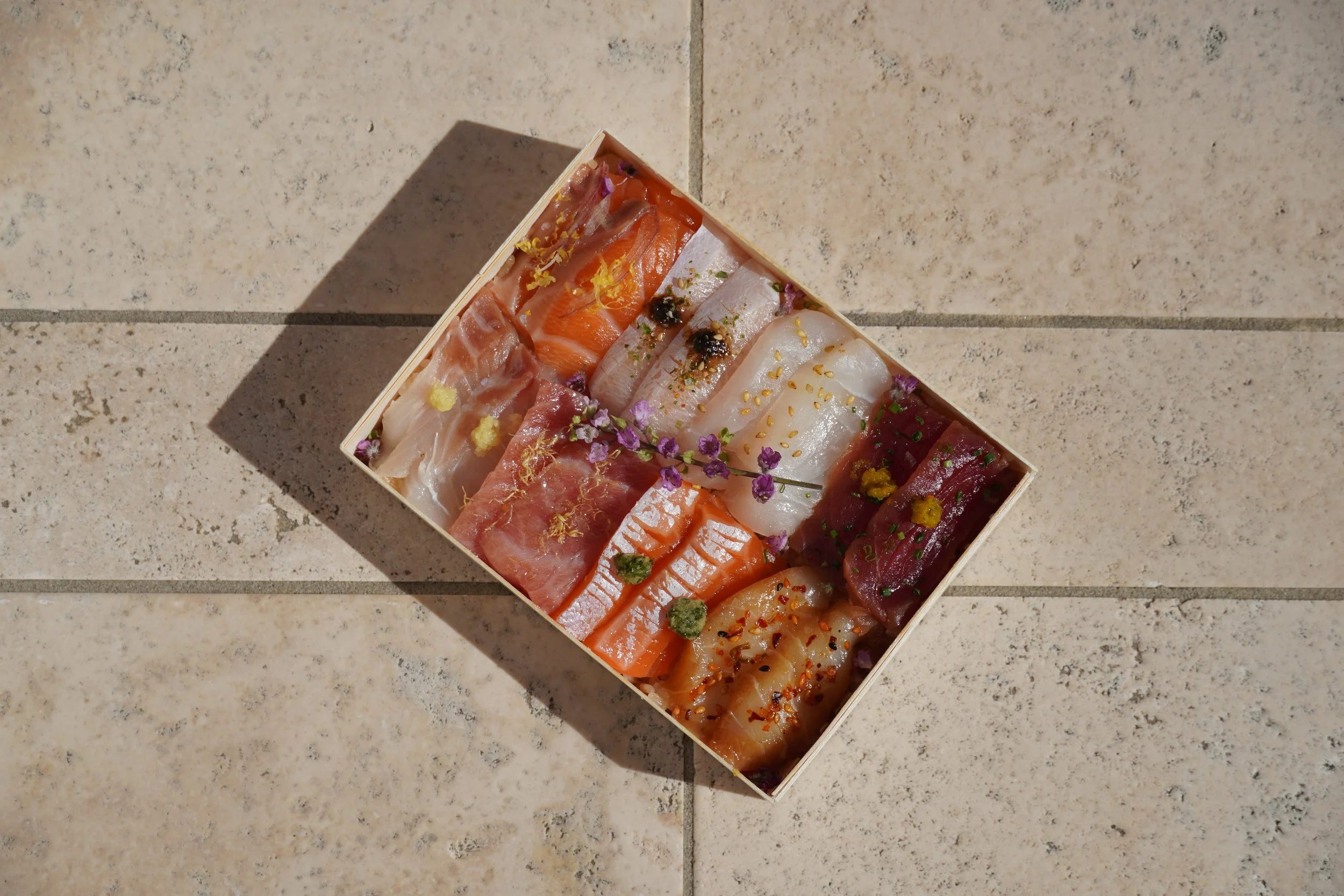 A rectangular box of assorted sashimi, including slices of fish such as salmon, tuna, shrimp, and white fish, garnished with edible flowers, wasabi, and sprinkled with spices, placed on a tiled surface.