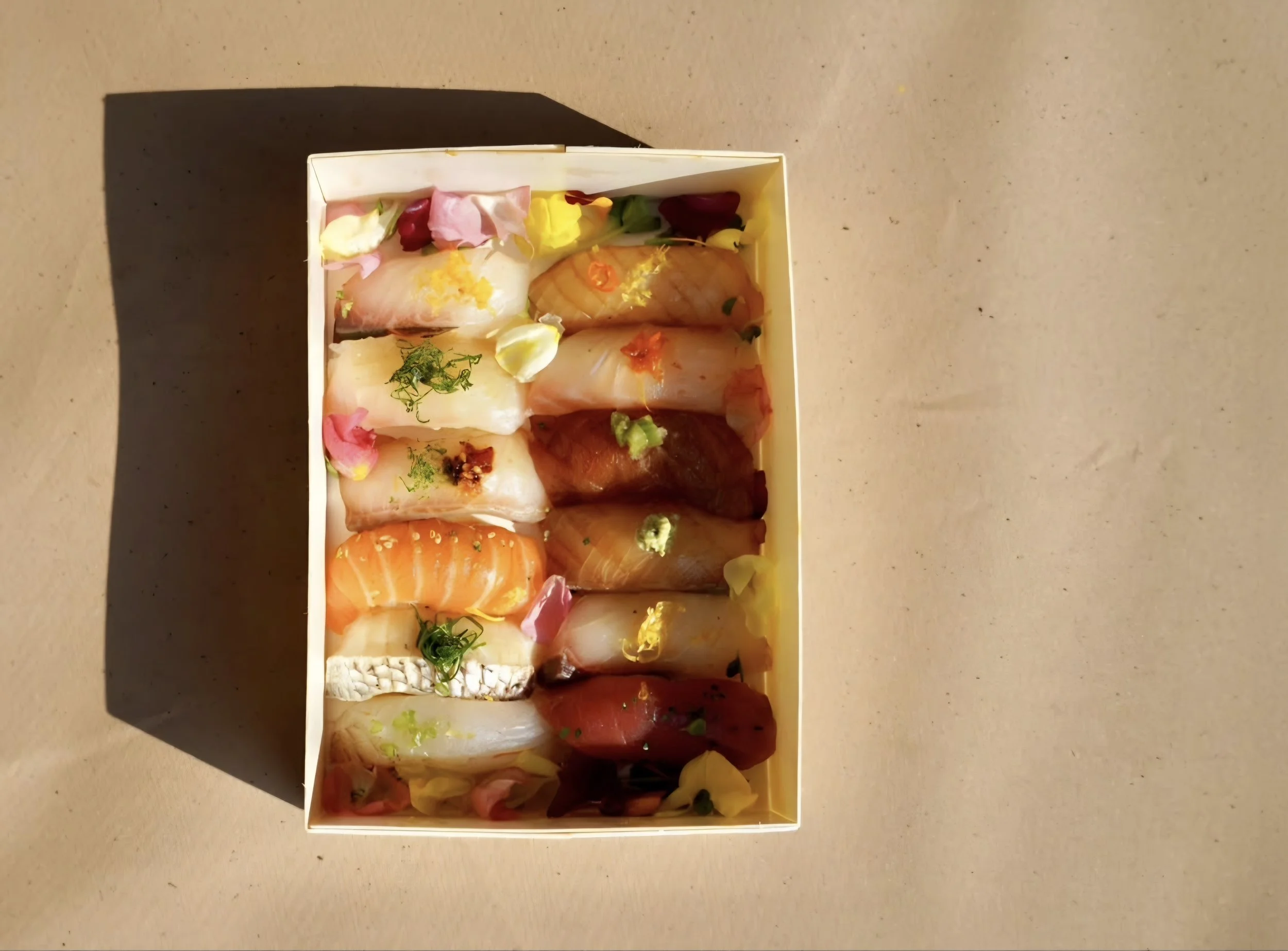 A white rectangular box filled with assorted pieces of sashimi, garnished with green herbs and edible flowers, placed on a beige table.