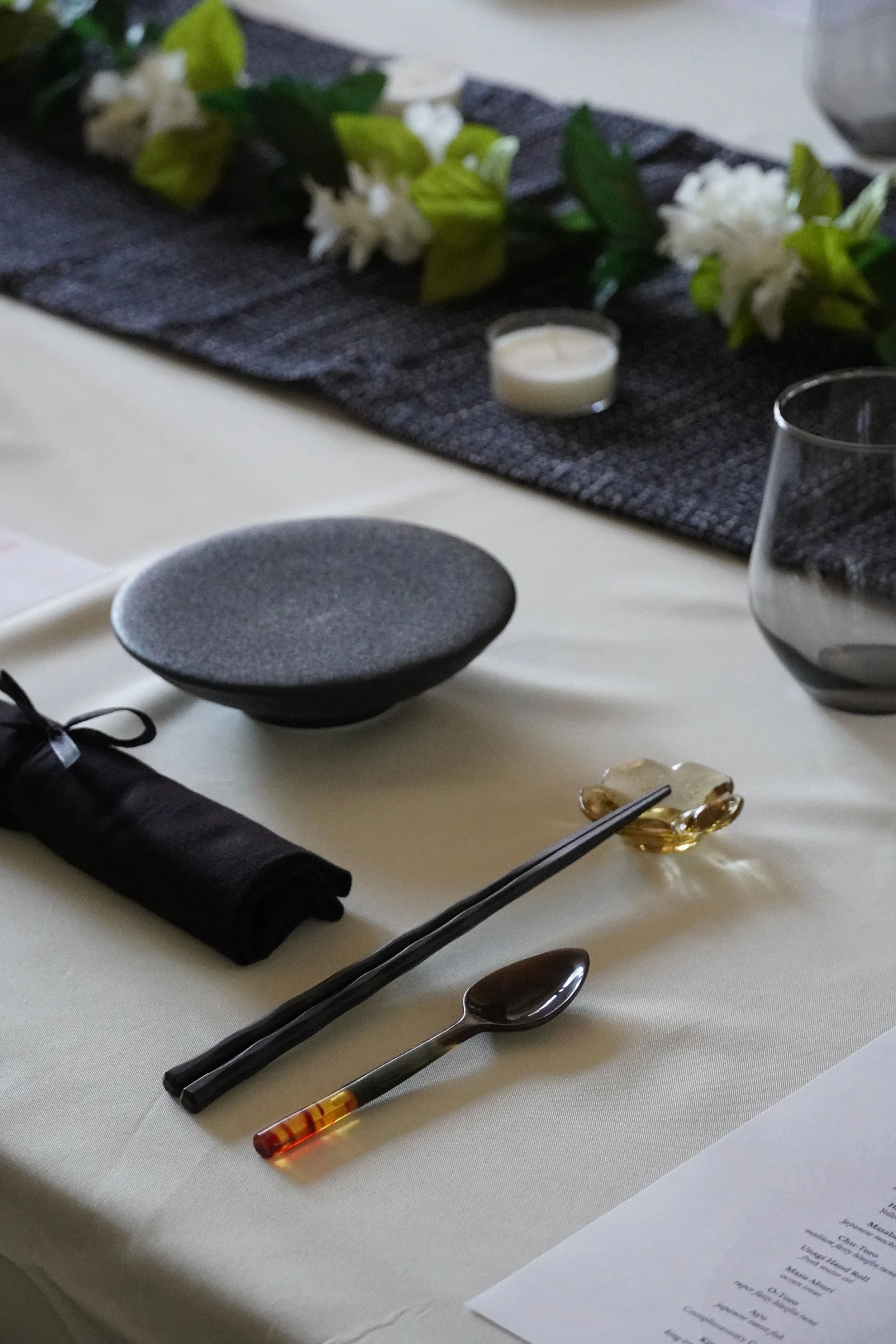 A close-up of a table set with black chopsticks, a spoon, a black rolled napkin, a small bowl of sauce, and a menu, with a decorative floral arrangement in the background.