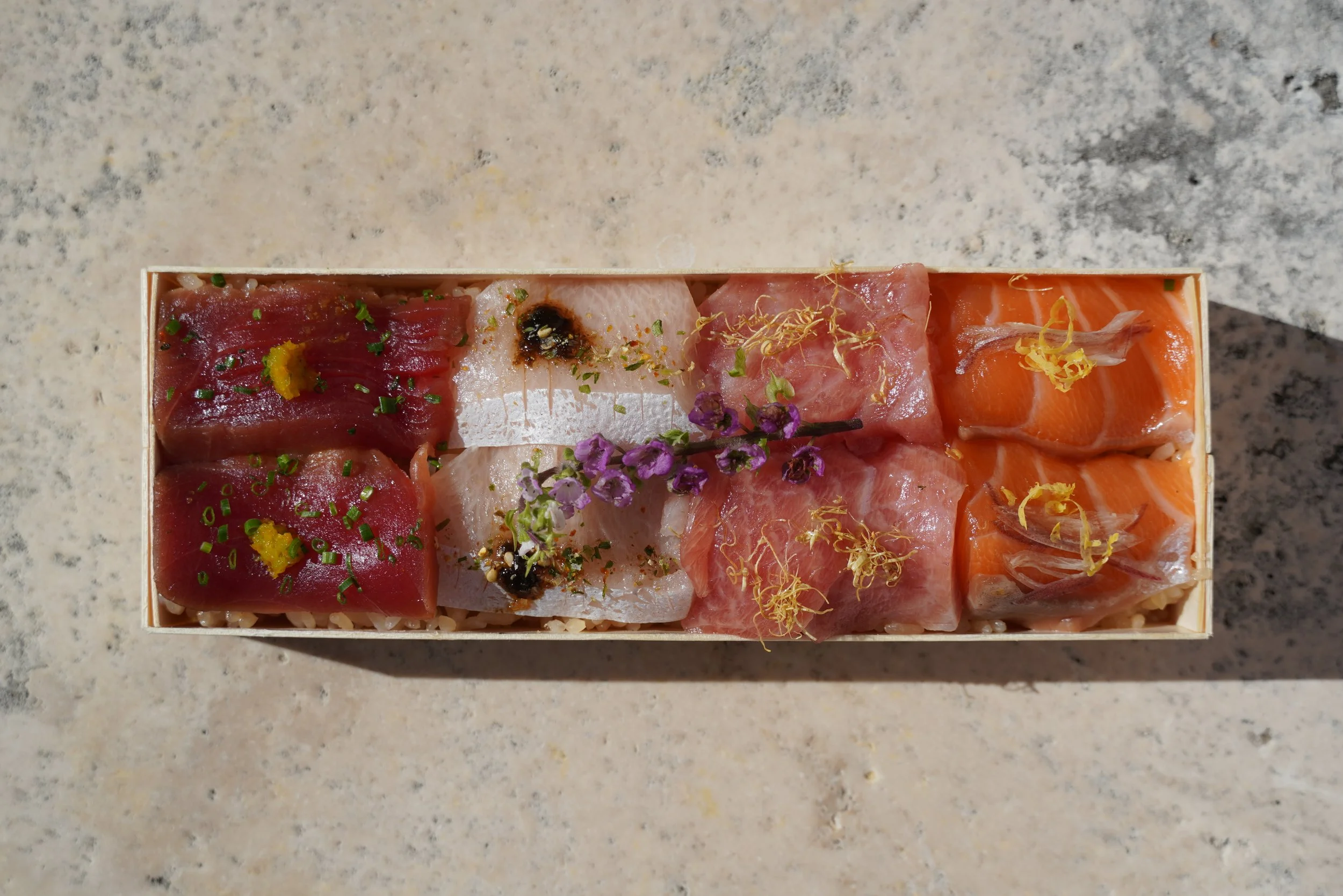 Assorted slices of sashimi including tuna, salmon, and white fish, garnished with microgreens, edible flowers, and citrus zest, arranged in a box on a speckled countertop.