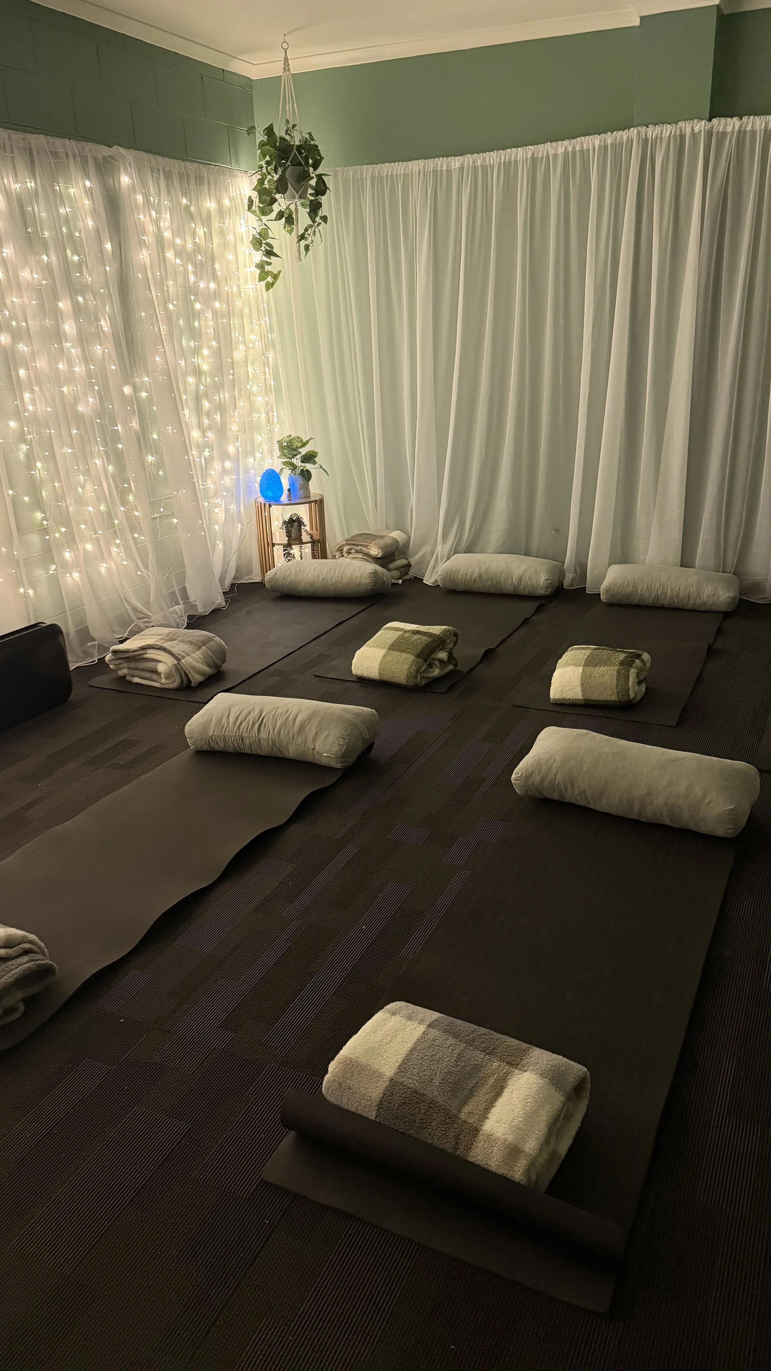 A cozy meditation or yoga space with black mats and rolled-up blankets arranged on top. The room has white curtains, some illuminated with string lights, green walls, and potted plants, including a hanging plant. A small side table holds a blue diffuser and a potted plant, creating a calming atmosphere.