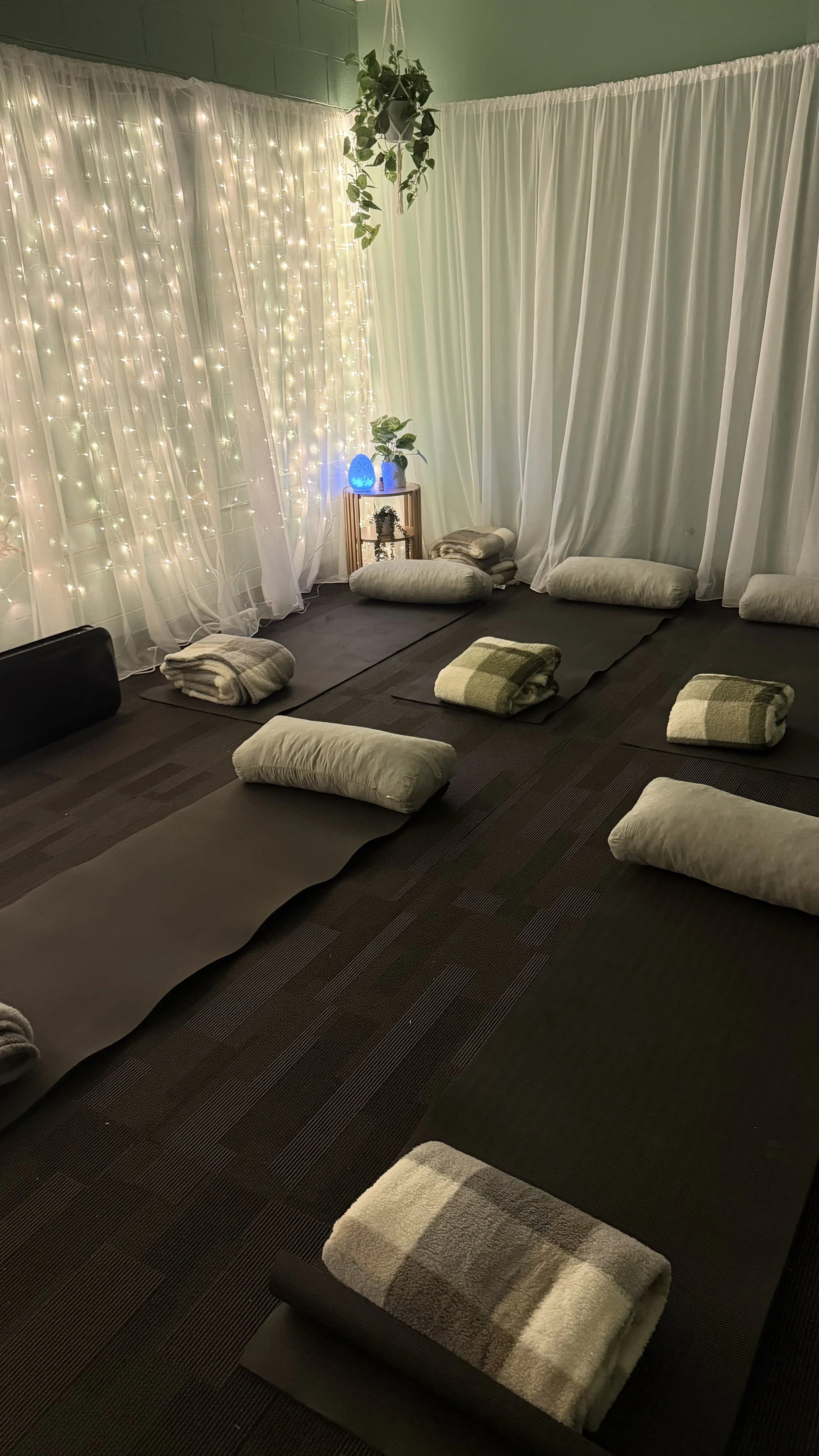 A cozy meditation or relaxation space with dark mats and rolled-up towels, cushioned pillows, and a small table with a salt lamp, plants, and a string of fairy lights behind sheer curtains.