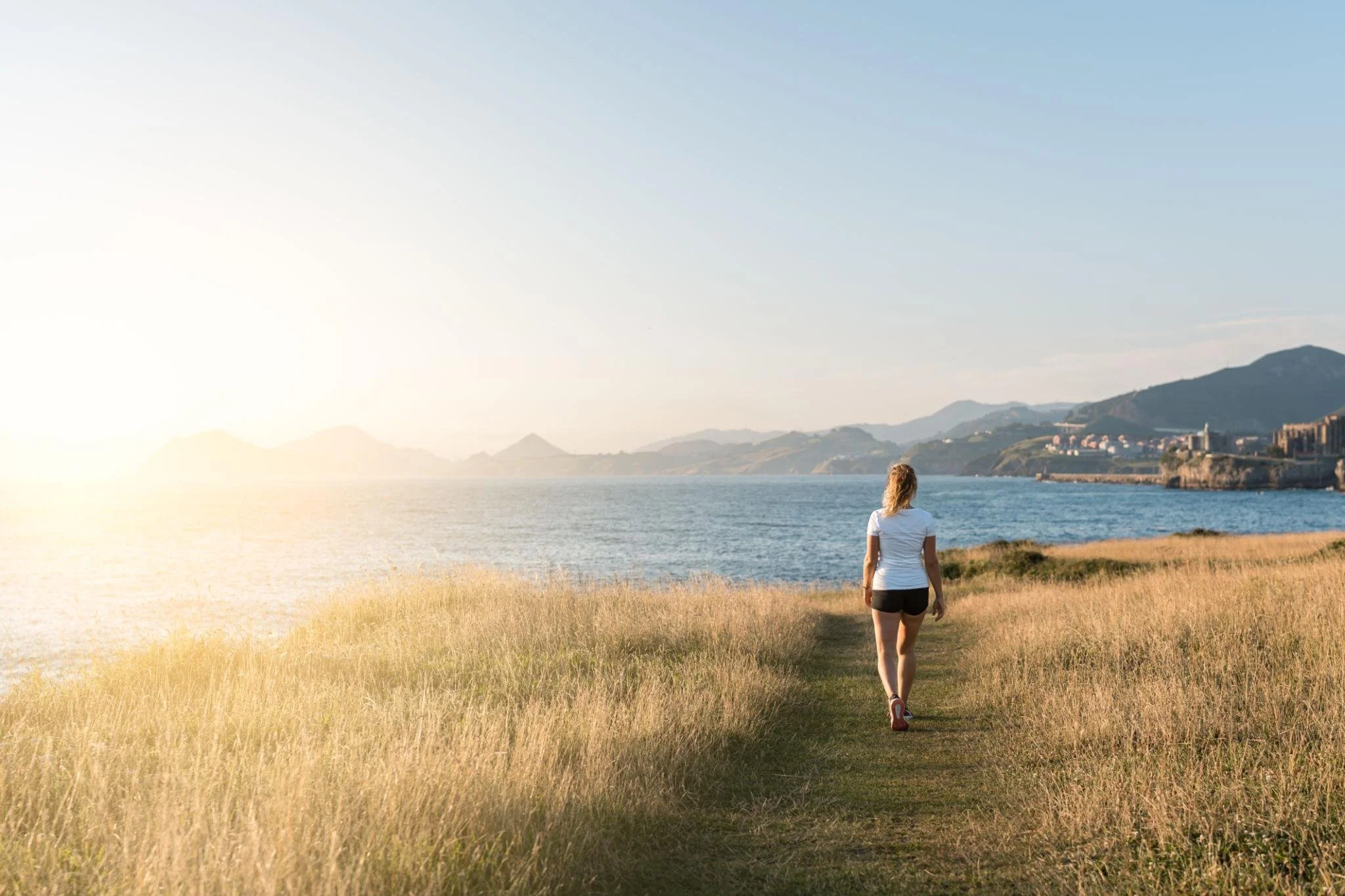 A woman walking along a grassy path by the water, overlooking distant mountains and buildings at sunset.