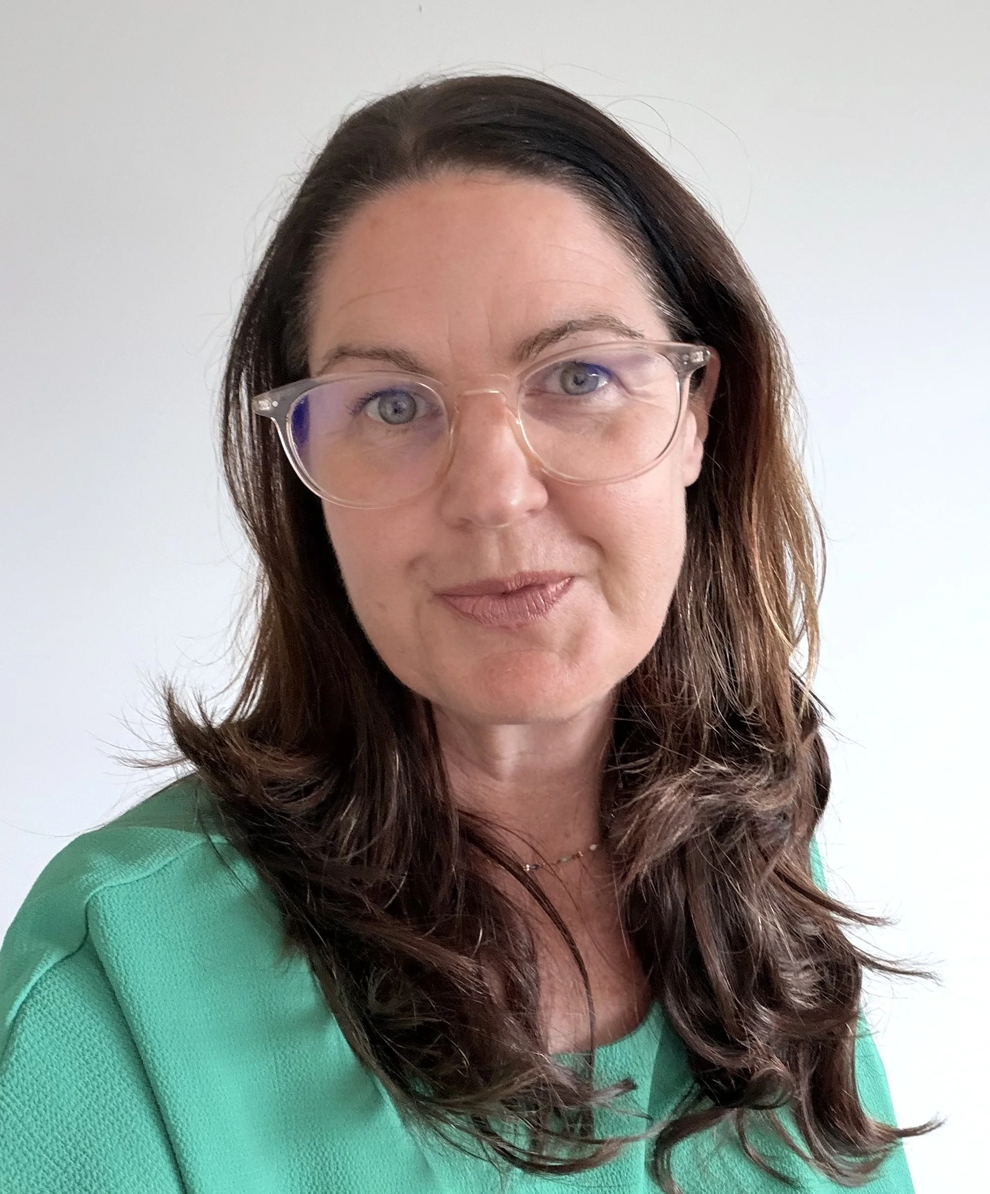A woman with long brown hair, wearing a green top and clear eyeglasses, smiling softly against a plain white background.