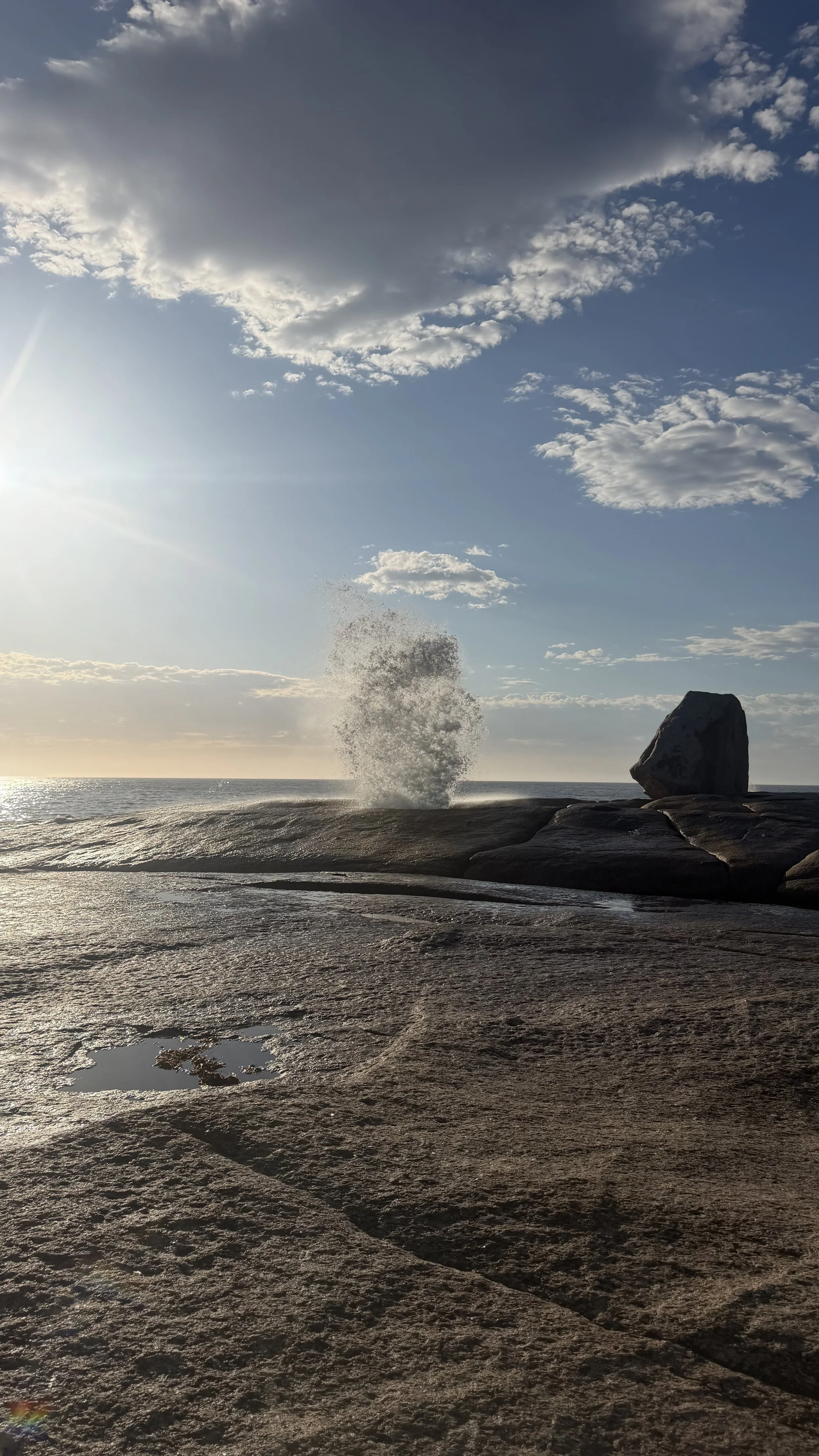 A rocky shoreline with a large wave crashing against a rock, with the ocean and a partly cloudy sky in the background, during sunset or sunrise.