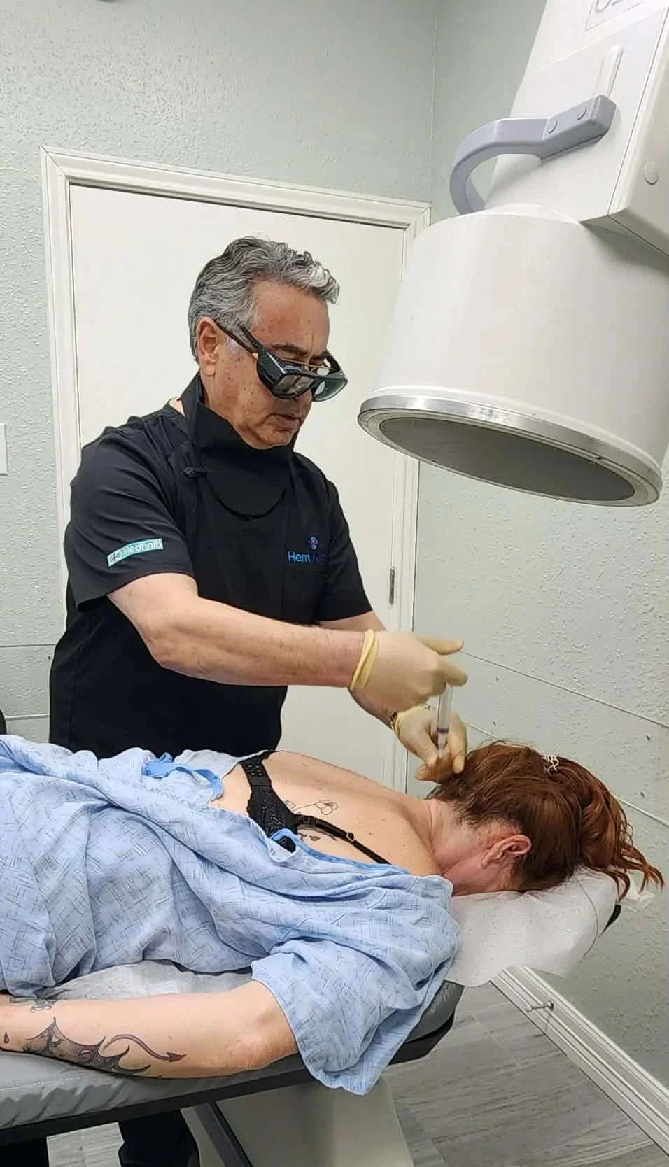 A doctor performing a tattoo removal procedure on a woman lying on a medical table in a clinical setting. The doctor is wearing protective glasses and gloves, and is using a handheld device to treat the woman's shoulder area.