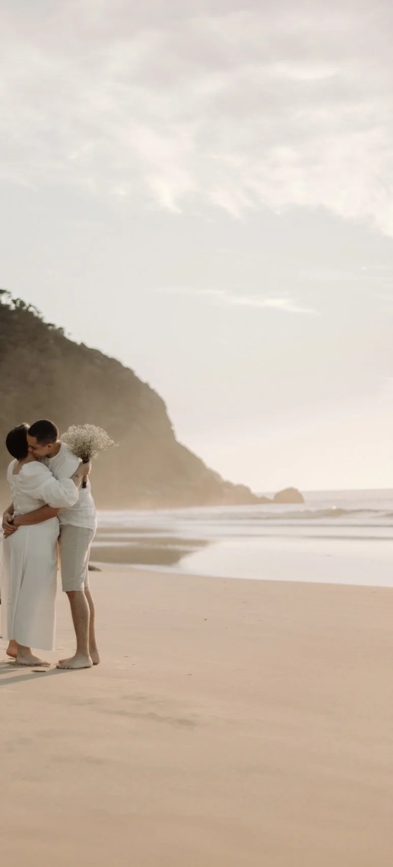A couple dressed in white clothes sharing a kiss on the beach during sunset, with a mountain and cloudy sky in the background.