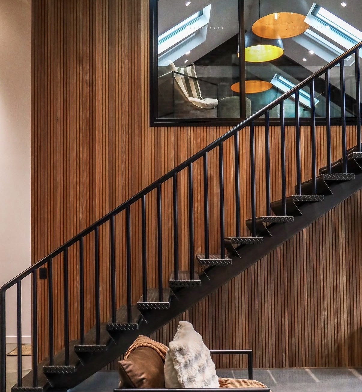 Interior of a modern building with black metal staircase, wood-paneled wall, and a window showing a cozy seating area with chairs and colorful round pendant lights.