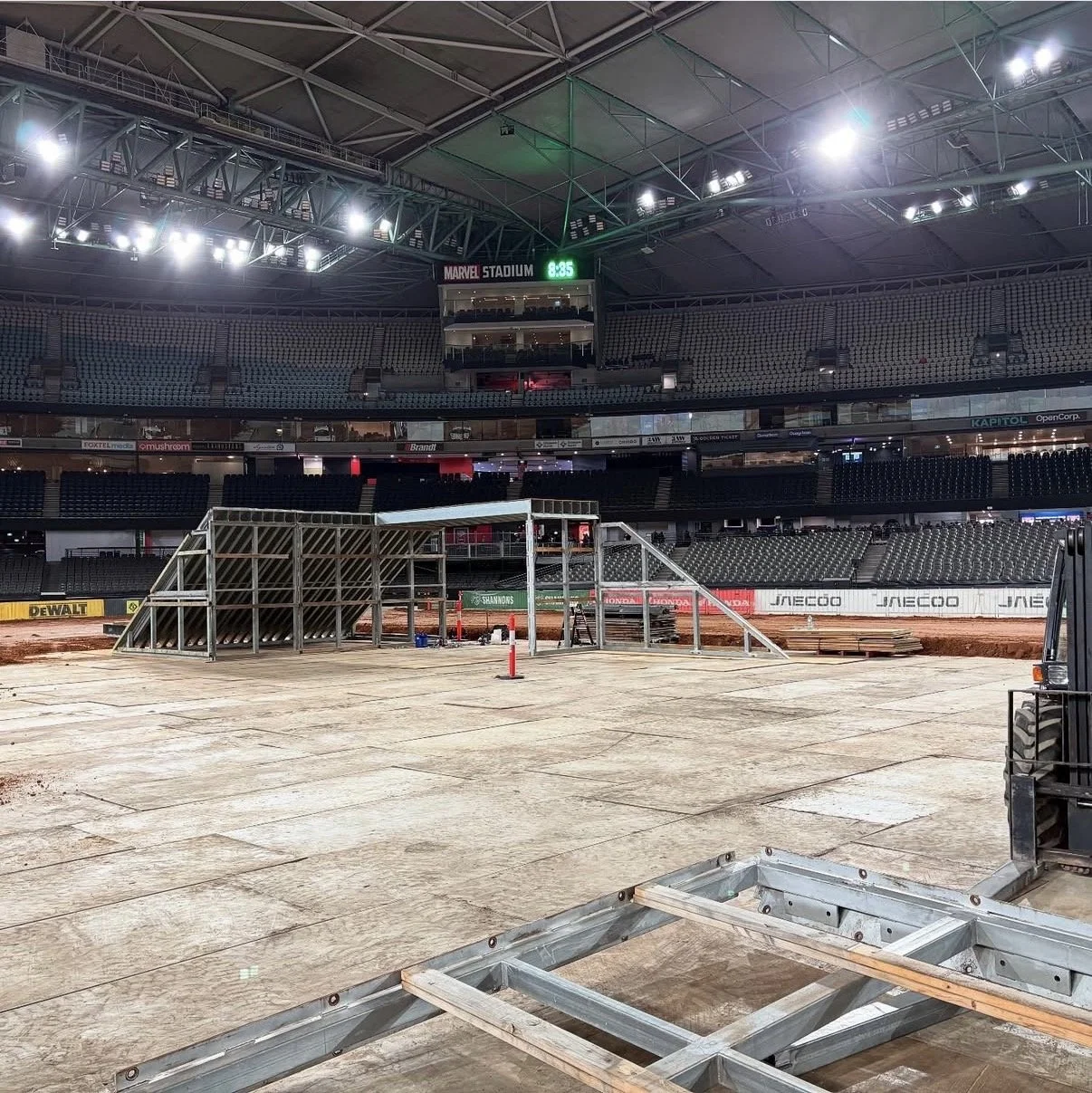 An indoor stadium under construction with metal framework and construction materials on the ground, surrounded by empty seating sections and a digital clock showing 8:35.