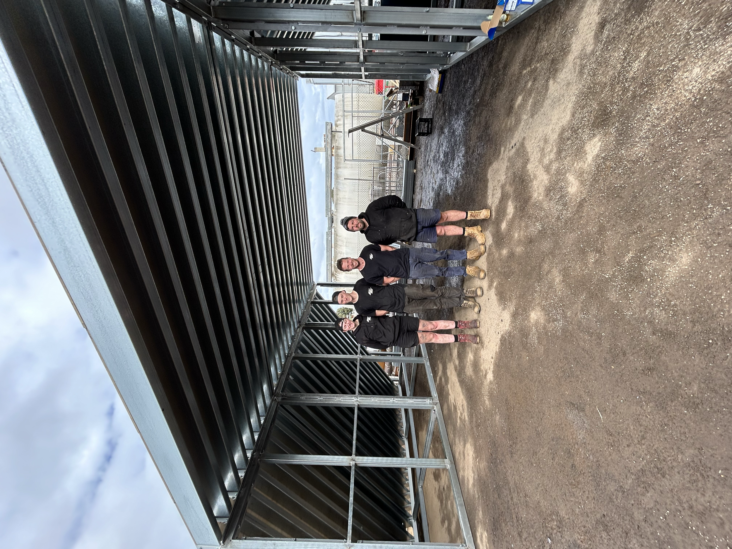 Four men standing under a metal roofing structure on a gravel ground near fencing, with machinery and cloudy sky in the background.