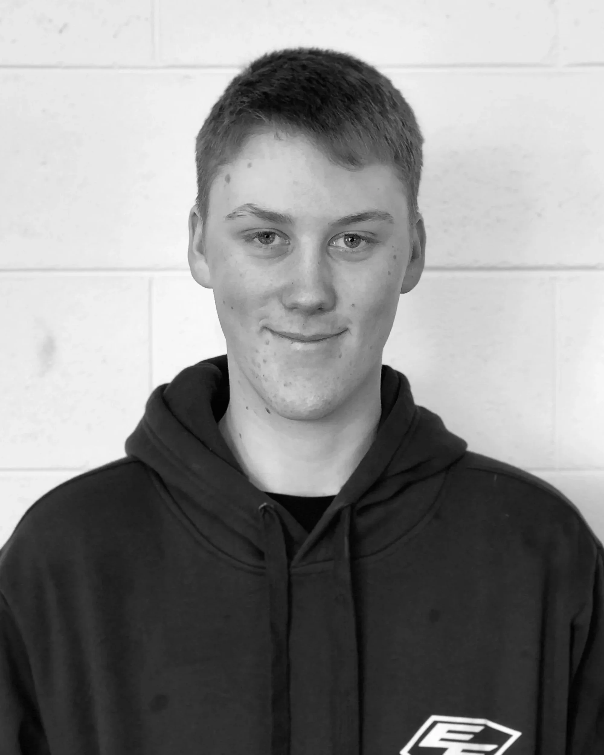 Close-up black-and-white photograph of a young man with short dark hair, wearing a black hoodie with a hexagon logo on the chest, standing against a plain white brick wall.