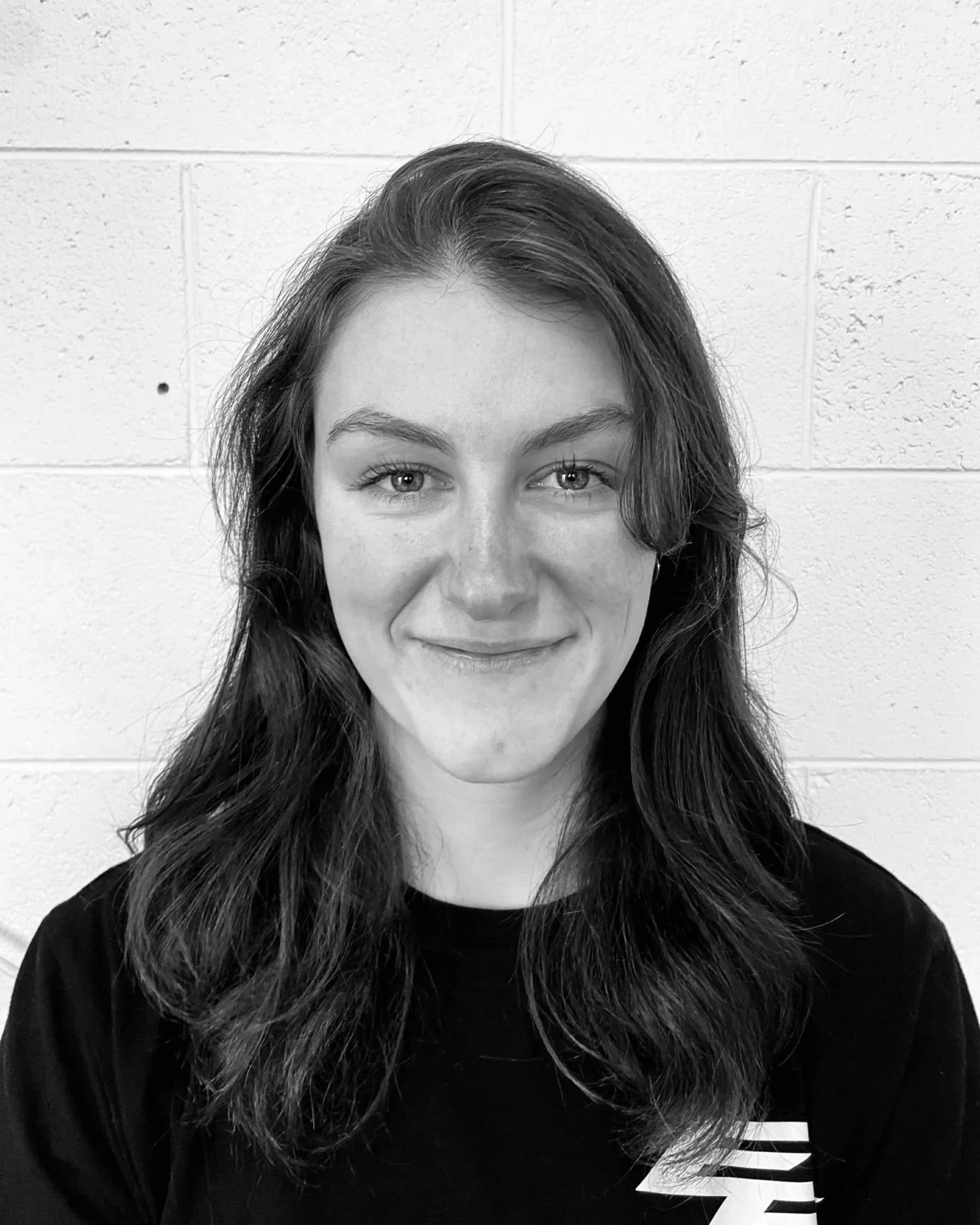 A black and white photo of a young woman with long, wavy hair, smiling softly, wearing a black top with a white striped sleeve, standing in front of a white, cinder block wall.