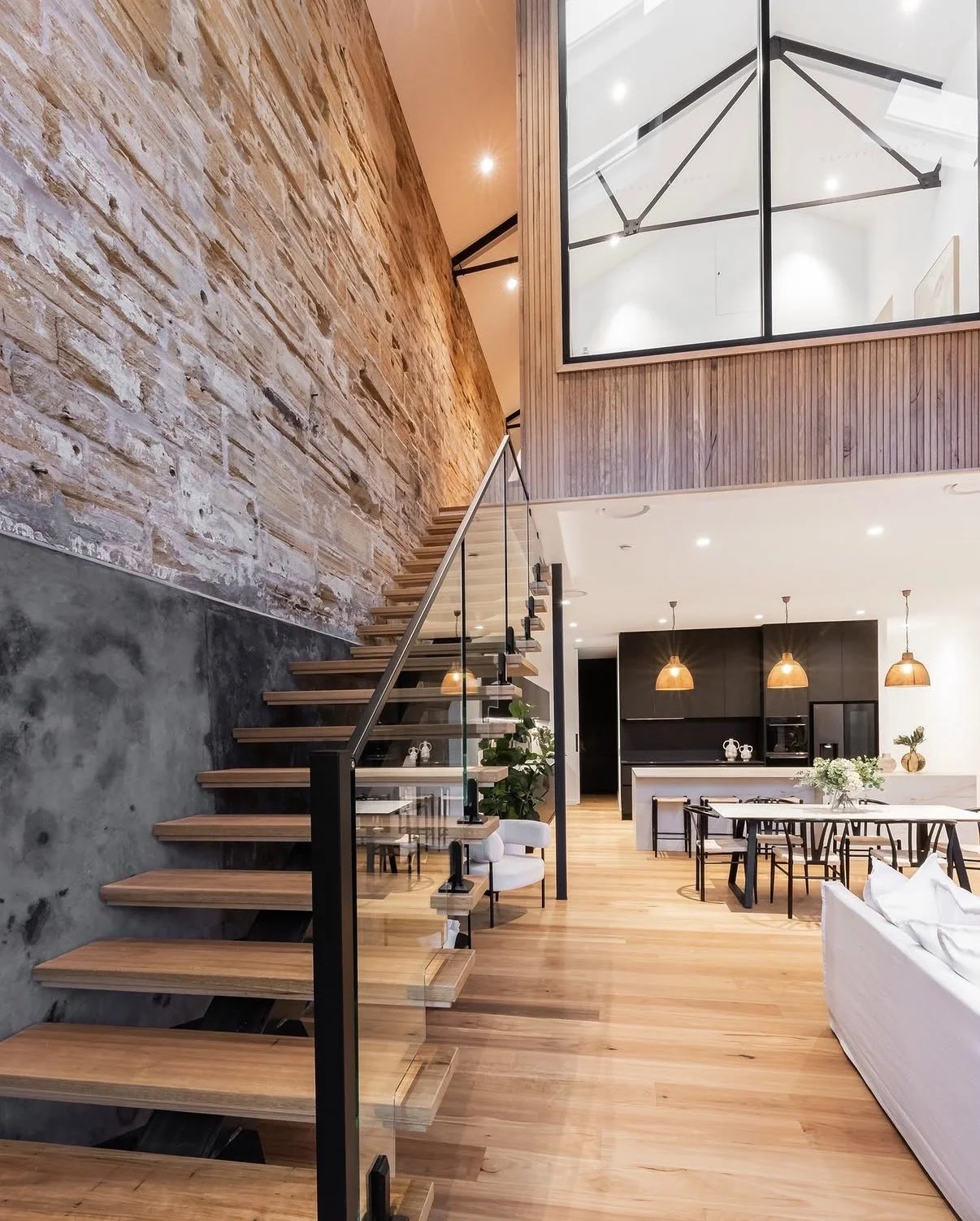 Modern open-concept living space with wooden stairs, exposed brick wall, black and white kitchen, and dining area with pendant lighting and floral centerpiece.
